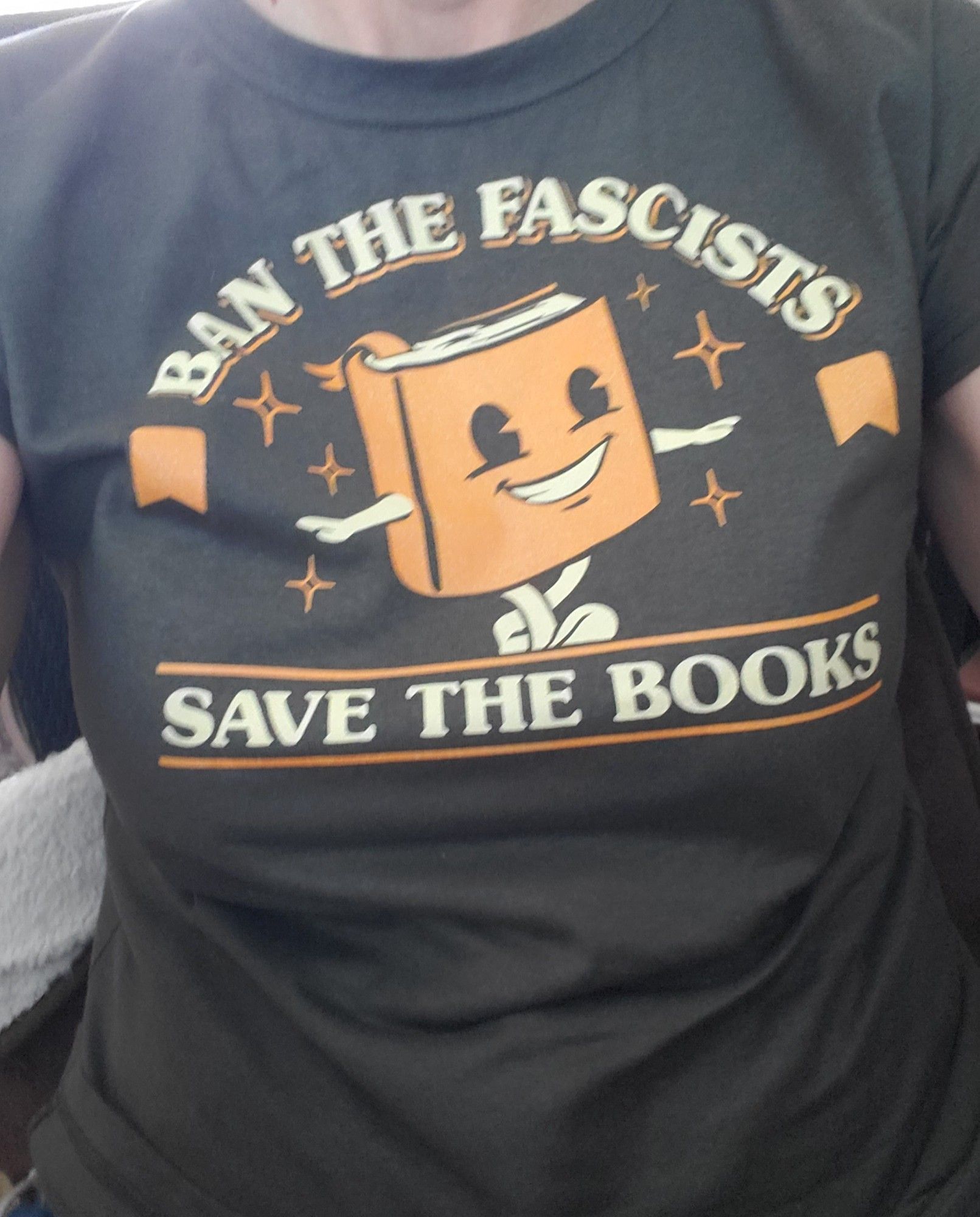 The front of a gray t-shirt which features a smiling cartoon book. Text reads "ban the fascists, save the books"