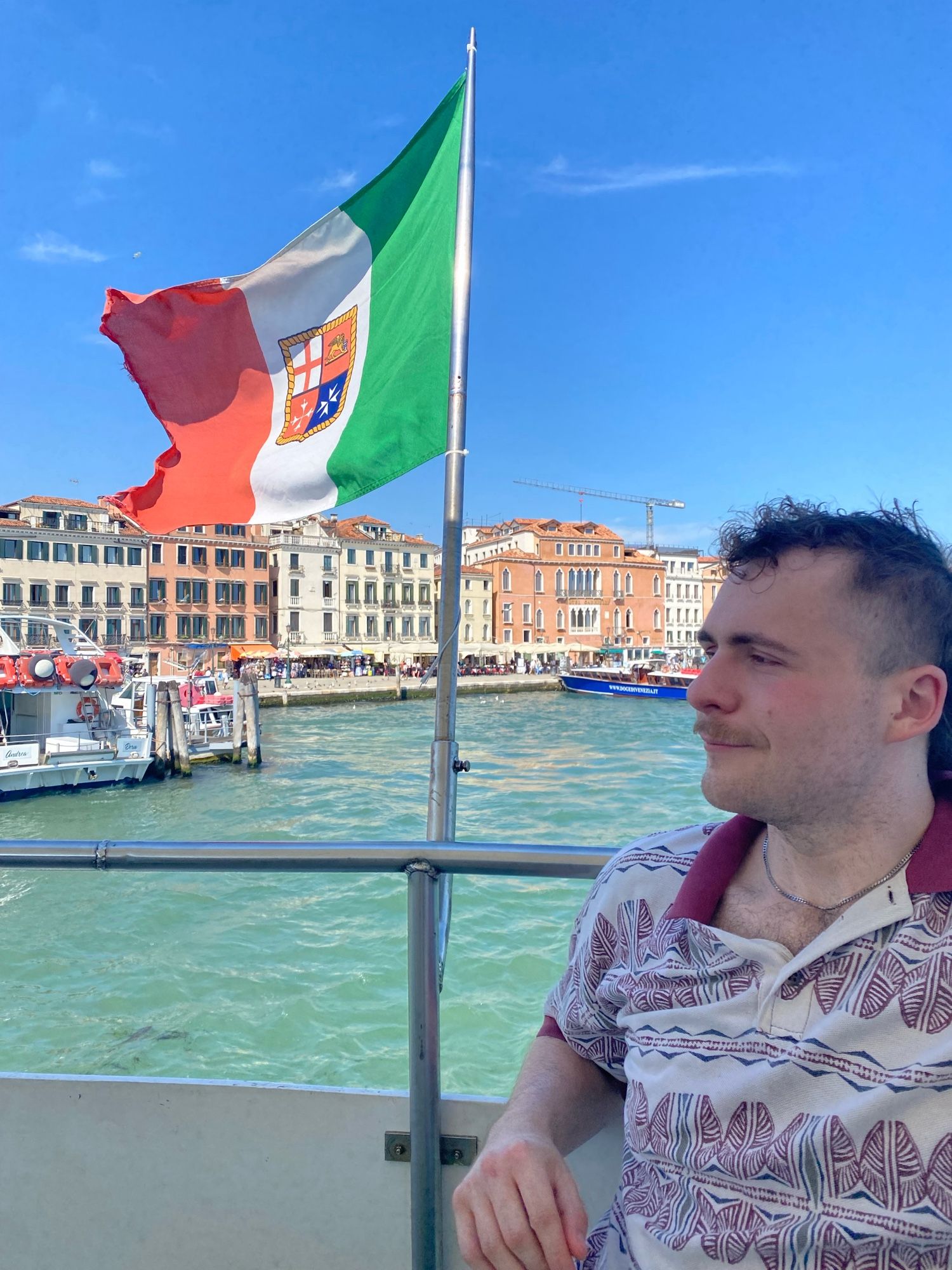 Aries looking off to the left on a boat, Venice in the background. The water is clear and there is a large Italian(? lol) flag flying in the middle of the photo