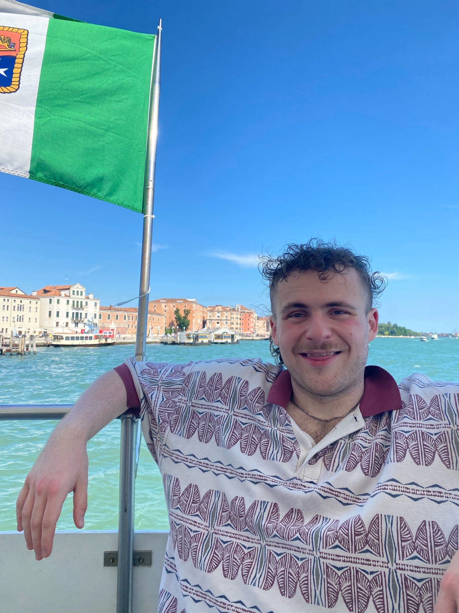 Aries cracking a goofy smile to the camera in a red and white patterned shirt , Venice in the background. The water is clear and so is the sky.