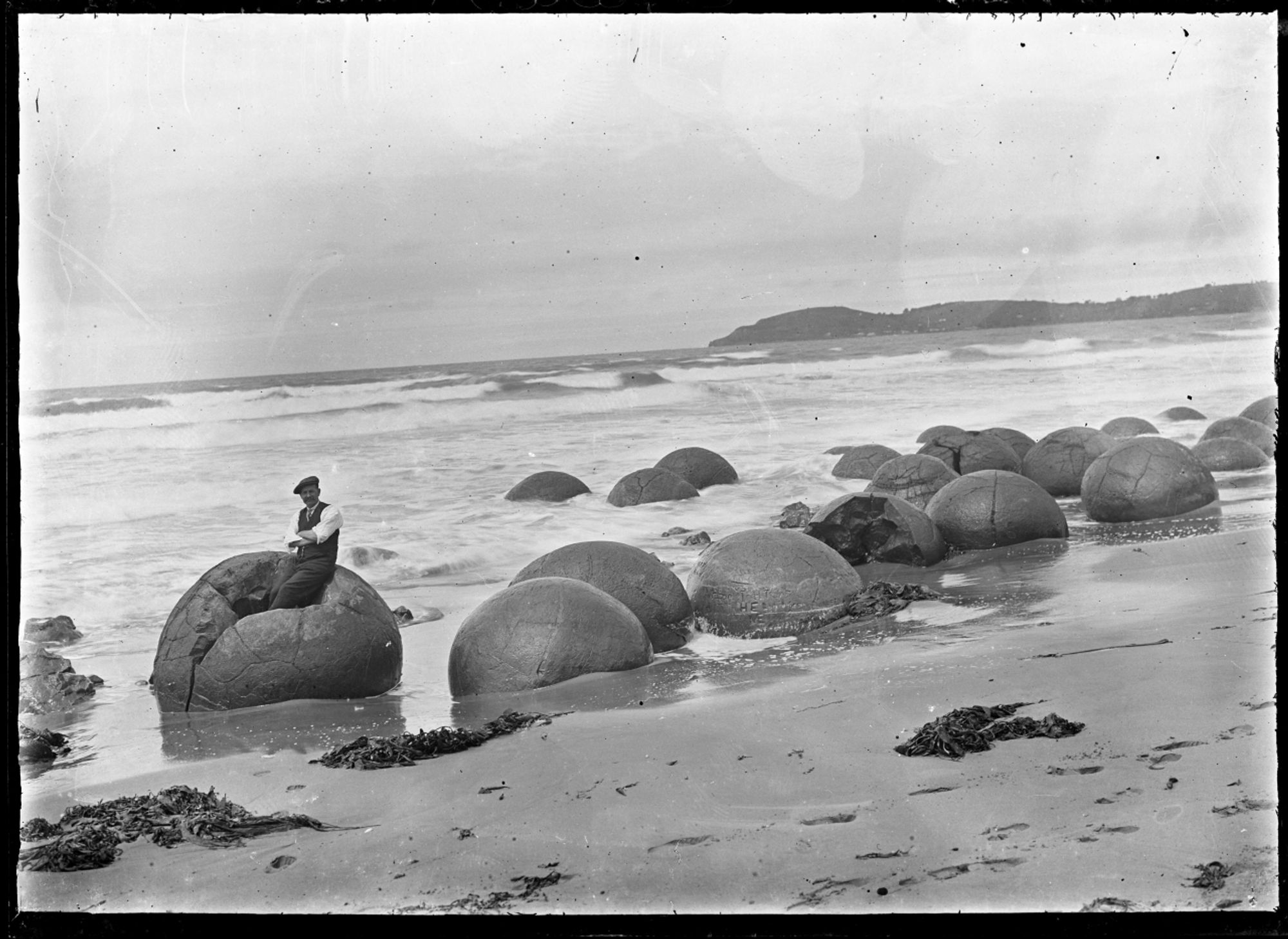 View of Moeraki Beach and boulders. A sense of scale is given to the boulders with the photographer Albert Percy Godber seated on one. Photograph taken by Albert Percy Godber circa 1925.