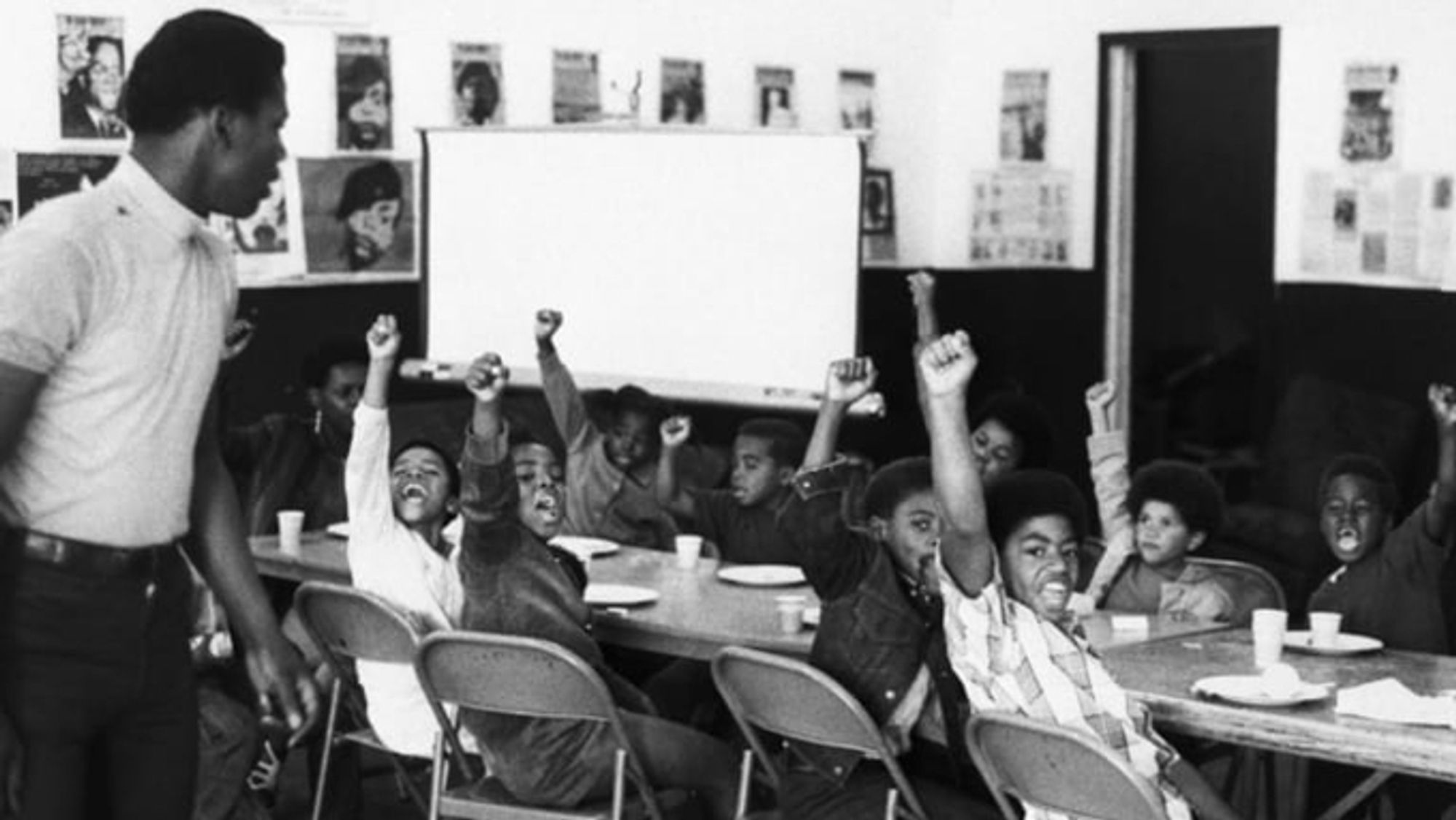 A well-dressed man in the foreground watches over a group of children sitting at a breakfast table with plates and cups.  The children have their fists raised and are shouting in unison.  The children and the man are Black, and the setting is the late 1960's.

BETTMANN ARCHIVE/GETTY IMAGES