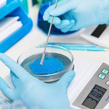 A pair of hands in a stock photo using a flat-ended spoon or scoop to transfer from a petri dish to a scientific weigh scale.  The substance is a granular blue powder.