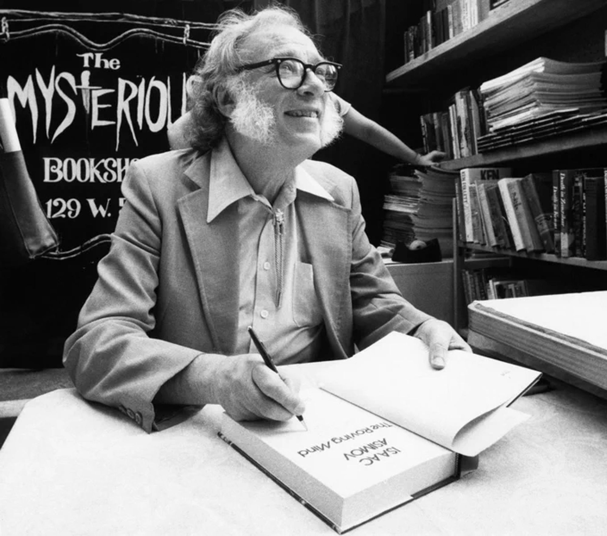 A man with dark-rim glasses signs a book in front of a storefront that reads "The Mysterious Bookshop".  He smiles, and is wearing a characteristic bolo tie, wide lapels, and tan suit.  He has mutton chops.

Isaac Asimov in 1984. (AP Photo/Mario Suriani)