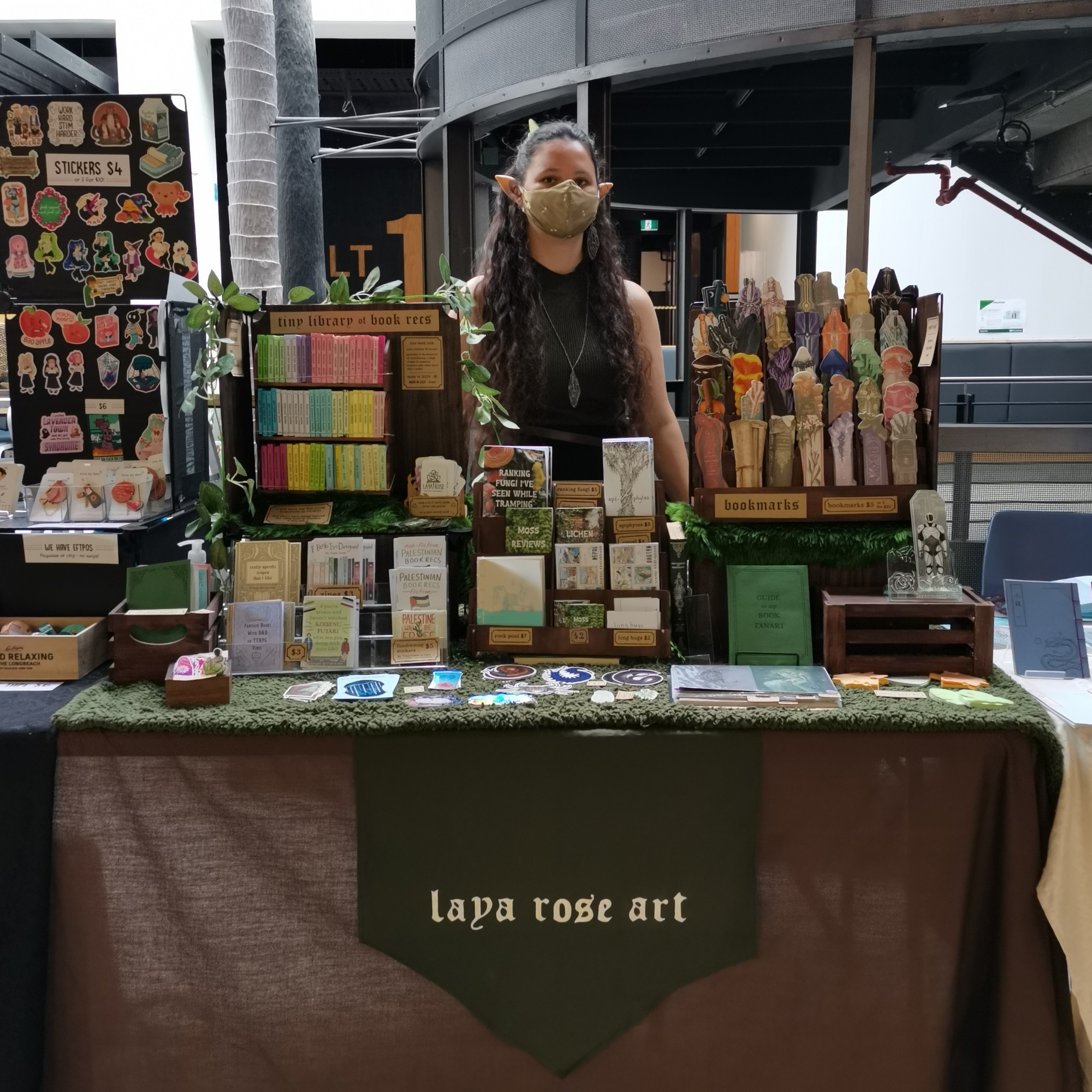 Photo of me at my artist alley stall. I have long hair, a Mask, and elf ears. My stall has various zines and bookmarks on a mossy tablecloth. There is a chevron banner that says Laya rose art. 