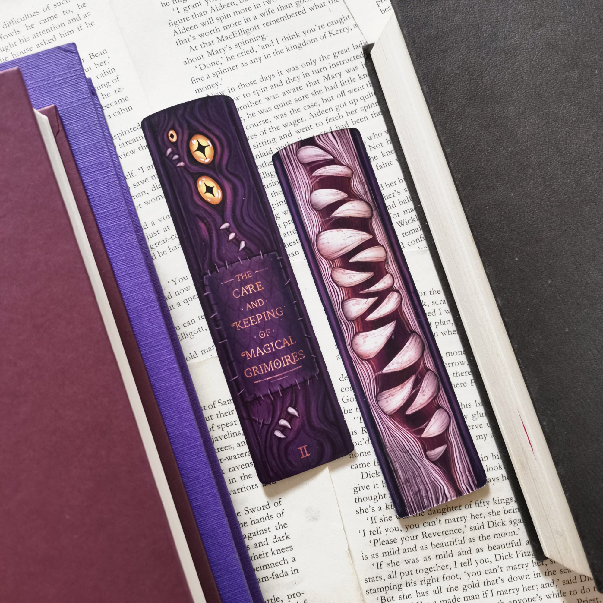 Photo of two sides of a monster themed bookmark with some purple hardcovers on the edges. The bookmark is a purple book spine that's stitched ridges leather with gold eyes and teeth, titled the care and keeping of magical grimoires. There's teeth on the page side.