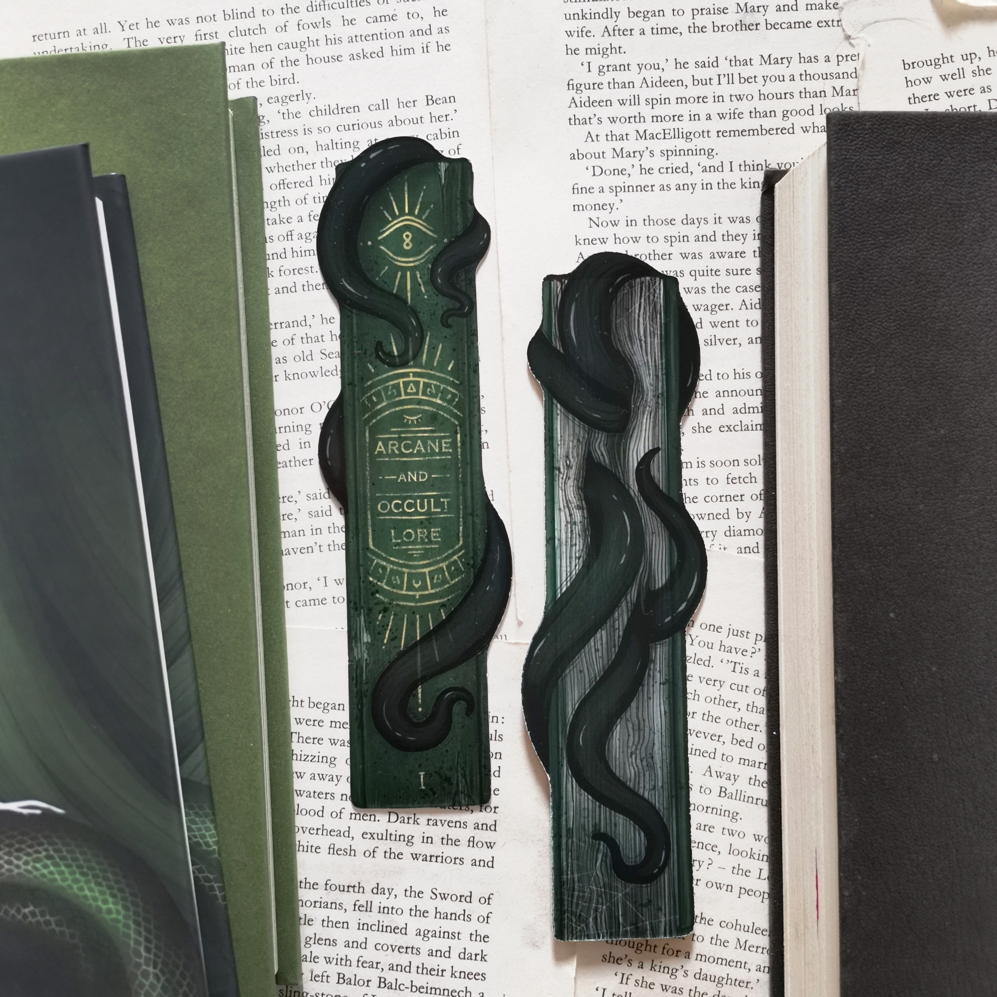 Photo of two sides of an eldritch themed bookmark with some green hardcovers on the edges. It's a green book spine titled arcane and occult lore, with some eye imagery and black tentacles coming out of it.