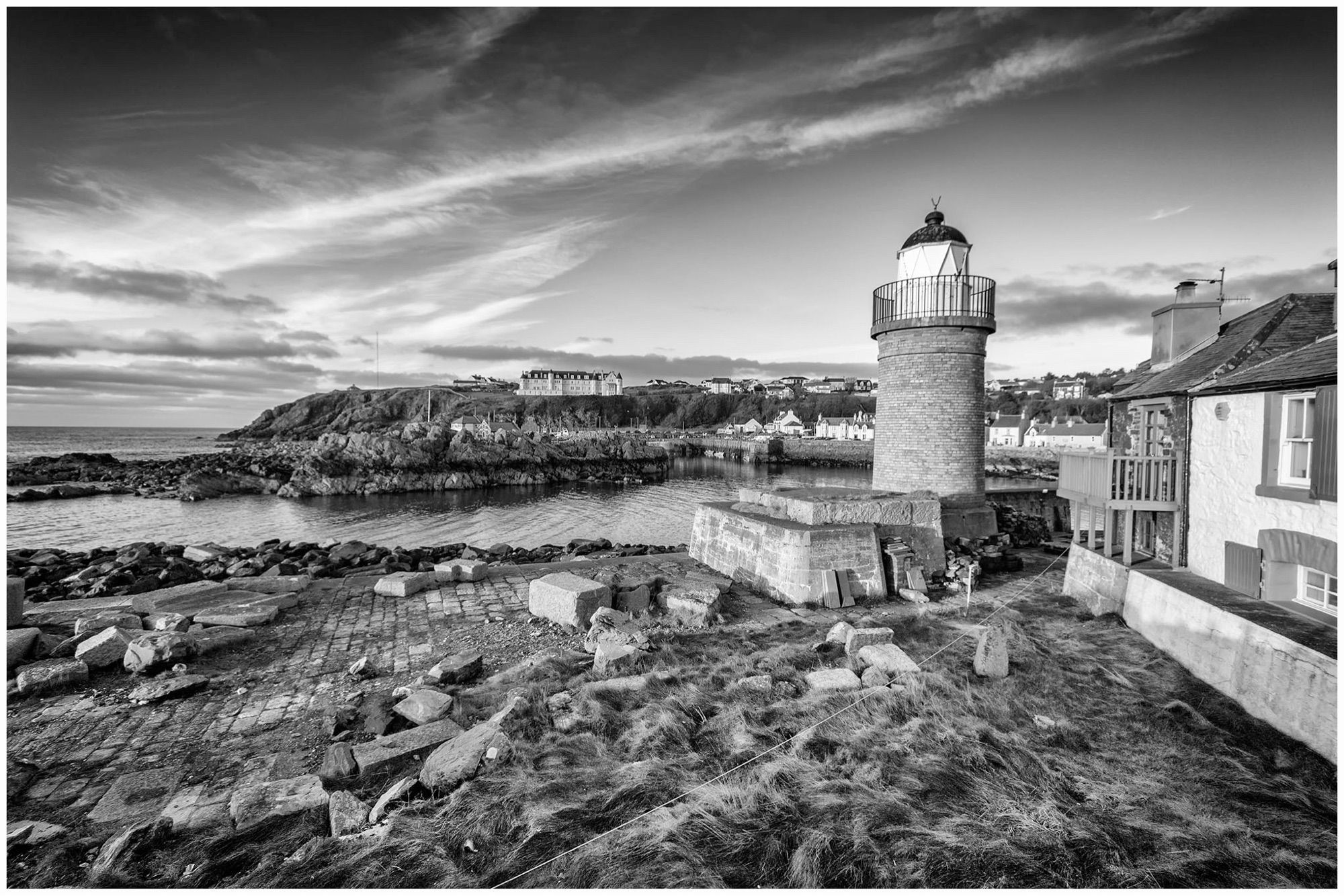 A black and white image of a coastal lighthouse beside a rocky shoreline. The lighthouse is on the right, with a small structure nearby. In the distance, a town is visible along the coast, with grassy areas and cloudy skies overhead.