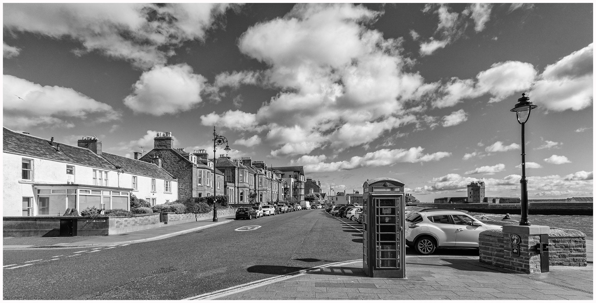 A black and white image depicting a coastal street with residential buildings on one side, parked cars, and a classic telephone booth. Fluffy clouds fill the sky, with a hint of sea visible in the background. A streetlamp and a sign can be