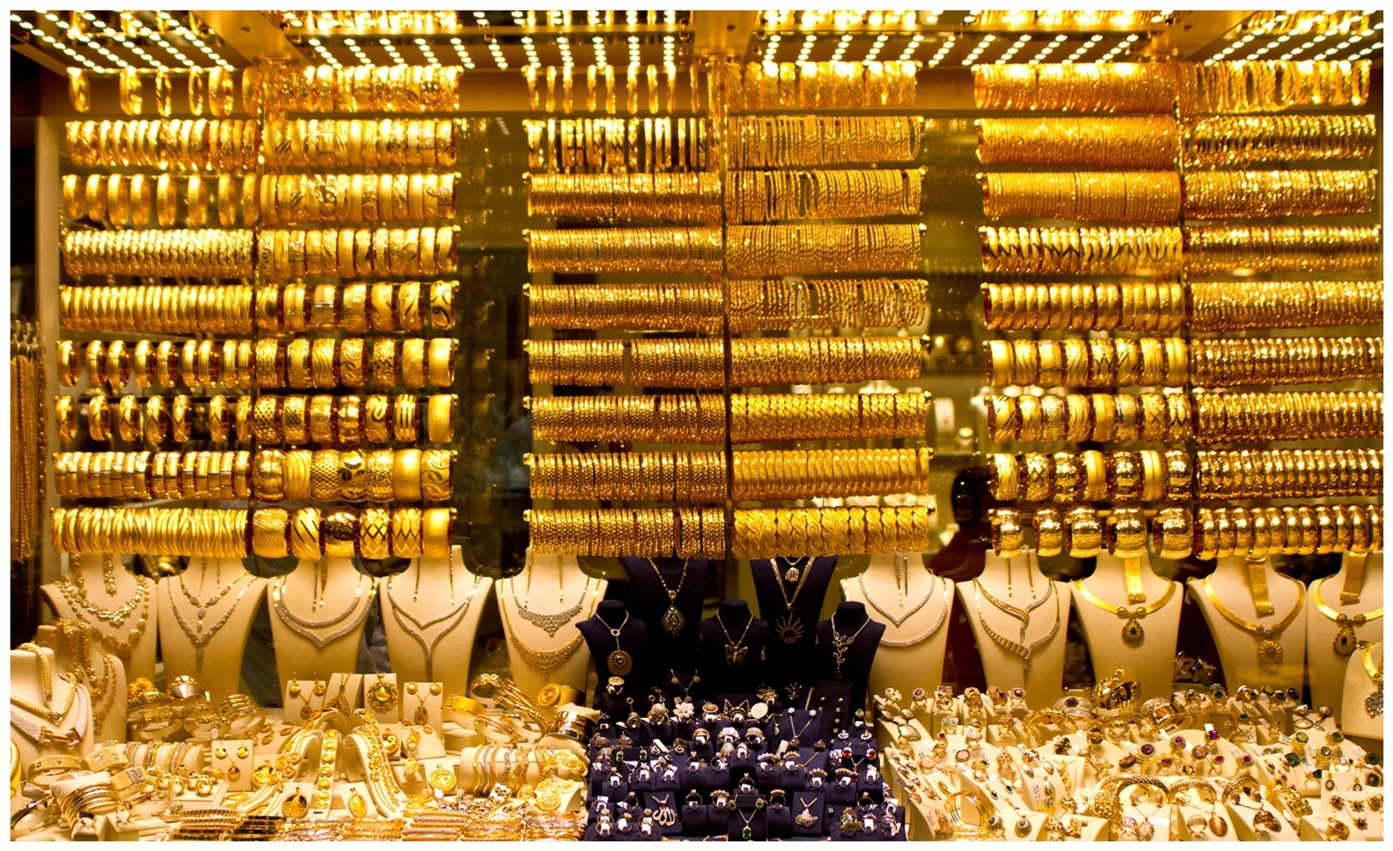 A display of gold jewelry including numerous gold bangles, necklaces, and rings arranged attractively in a window. The items are illuminated, highlighting their shine and intricate designs.