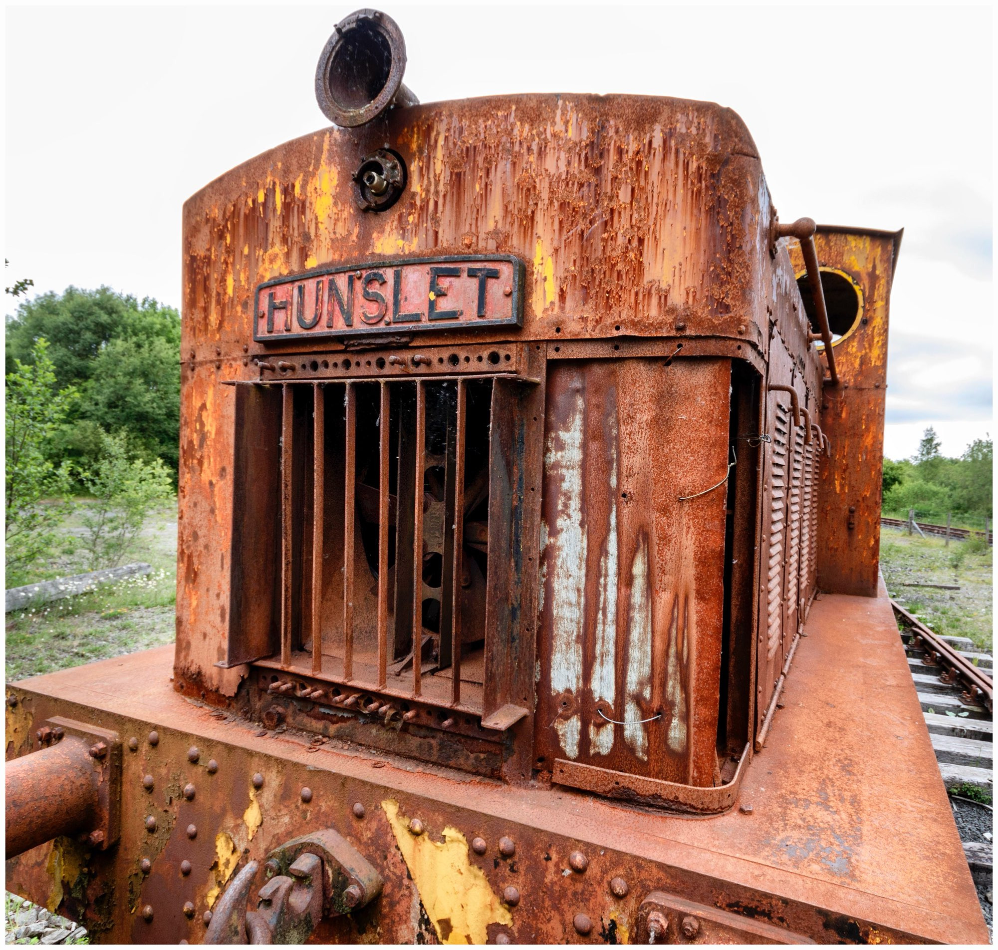 An old, rusty Hunslet locomotive is situated on overgrown tracks. The locomotive features a weathered exterior with significant corrosion and peeling paint, showcasing a vintage industrial aesthetic. Green vegetation surrounds the area, contrasting with the metal.