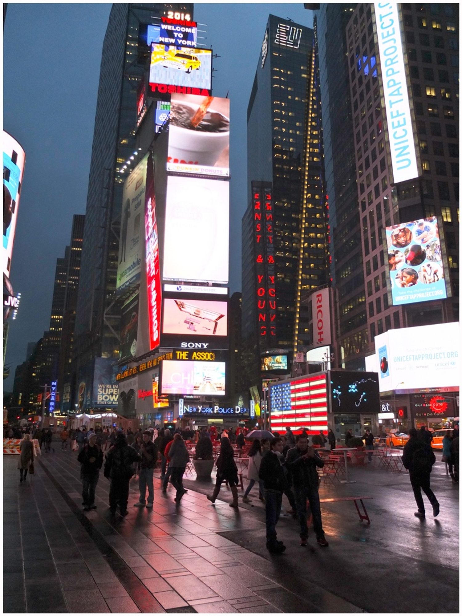 A bustling Times Square at night, featuring bright digital billboards, a large American flag display, and crowds of pedestrians walking along the wet street. The atmosphere is lively, illuminated by various advertisements.