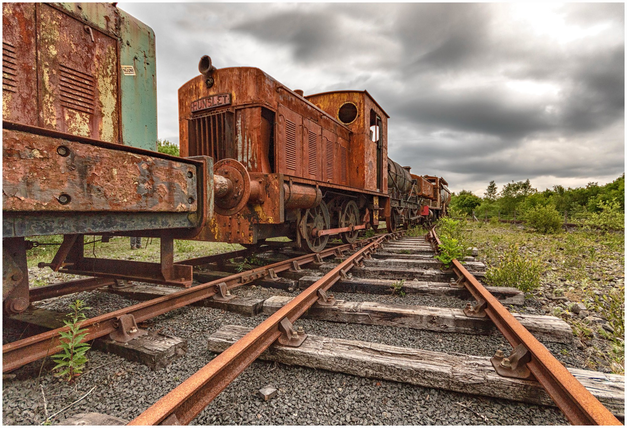 An abandoned rusted train on overgrown railway tracks, partially obscured by vegetation, under a cloudy sky.