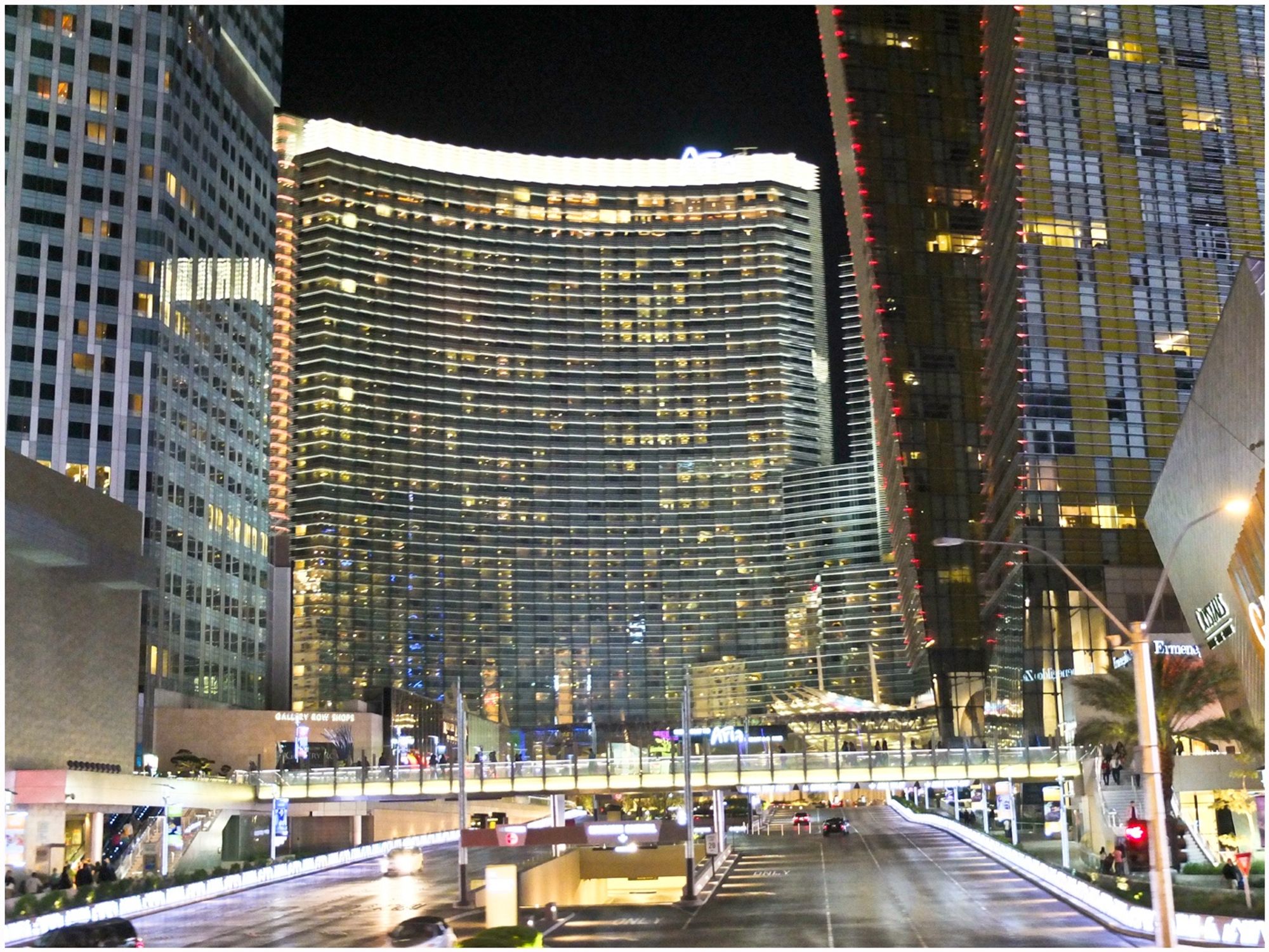 A cityscape at night featuring modern skyscrapers, including a curved building with illuminated edges. The scene includes a street with vehicle traffic and pedestrian walkways, showcasing a vibrant urban atmosphere.