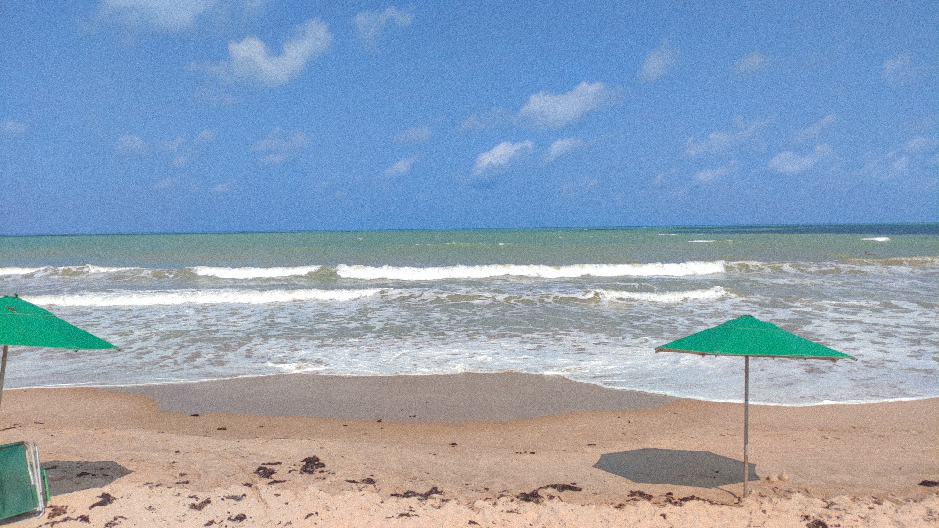 Foto da praia, tirada já da areia. Dá para ver três partes da areia: mais perto de onde tirei a foto, ela está seca e clara e fofa, logo depois está mais escura onde o mar acabou de retroceder, e logo após isso ainda claramente molhada e brilhanso onde a ágia ainda está sendo absorvida. Há duas sobrinhas, uma do lado direito, verde, e uma cortada do lado esquerdo. O mar está seco e tem pequenas ondas brancas, no fundo dá para ver ele verdinho, e o cêu super azul com poucas nuvens espalhadas