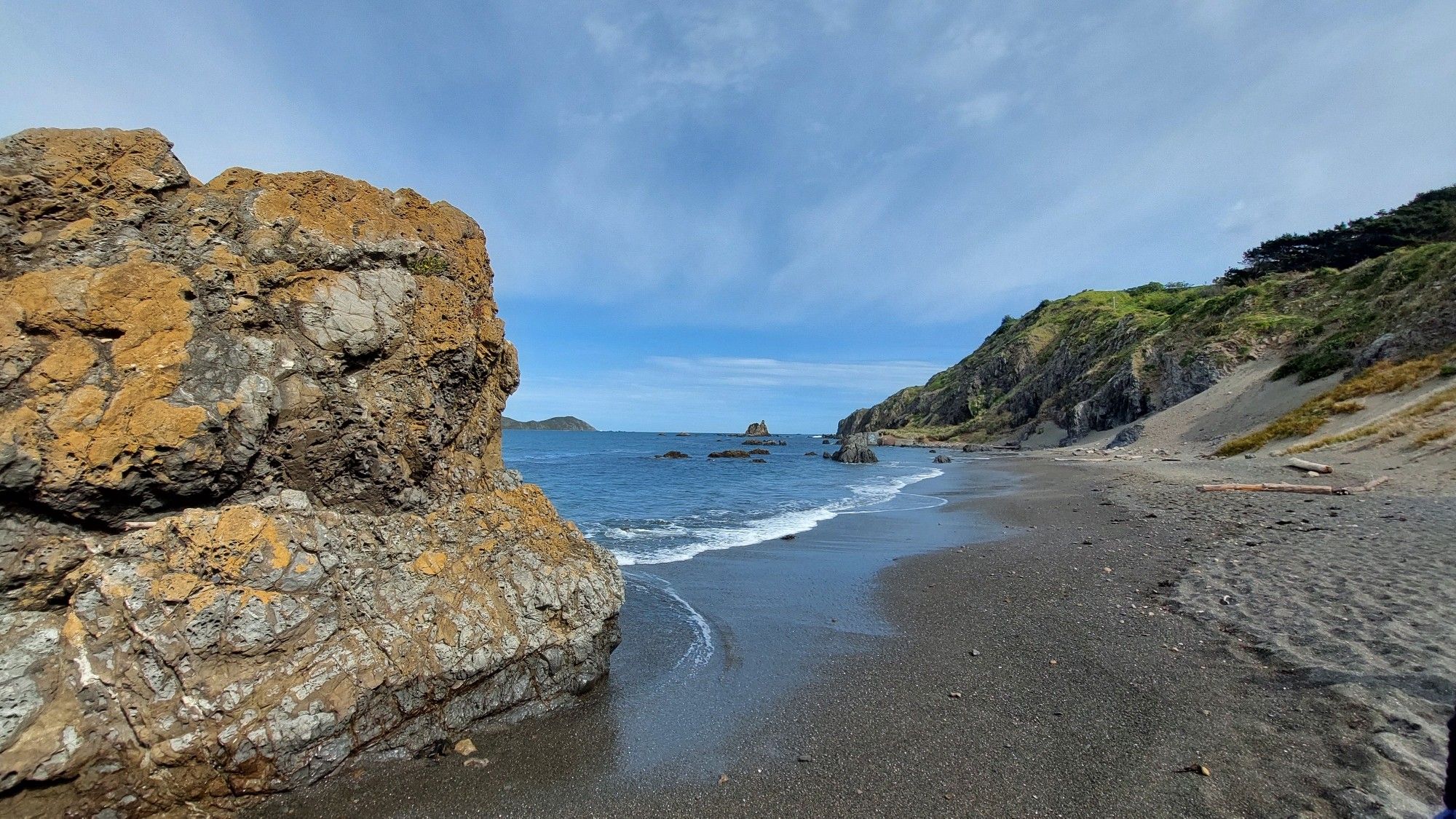 Brown rock sticking out of the water to the left and a stoney beach and cliff face on the right, blue skies and the ocean in the centre