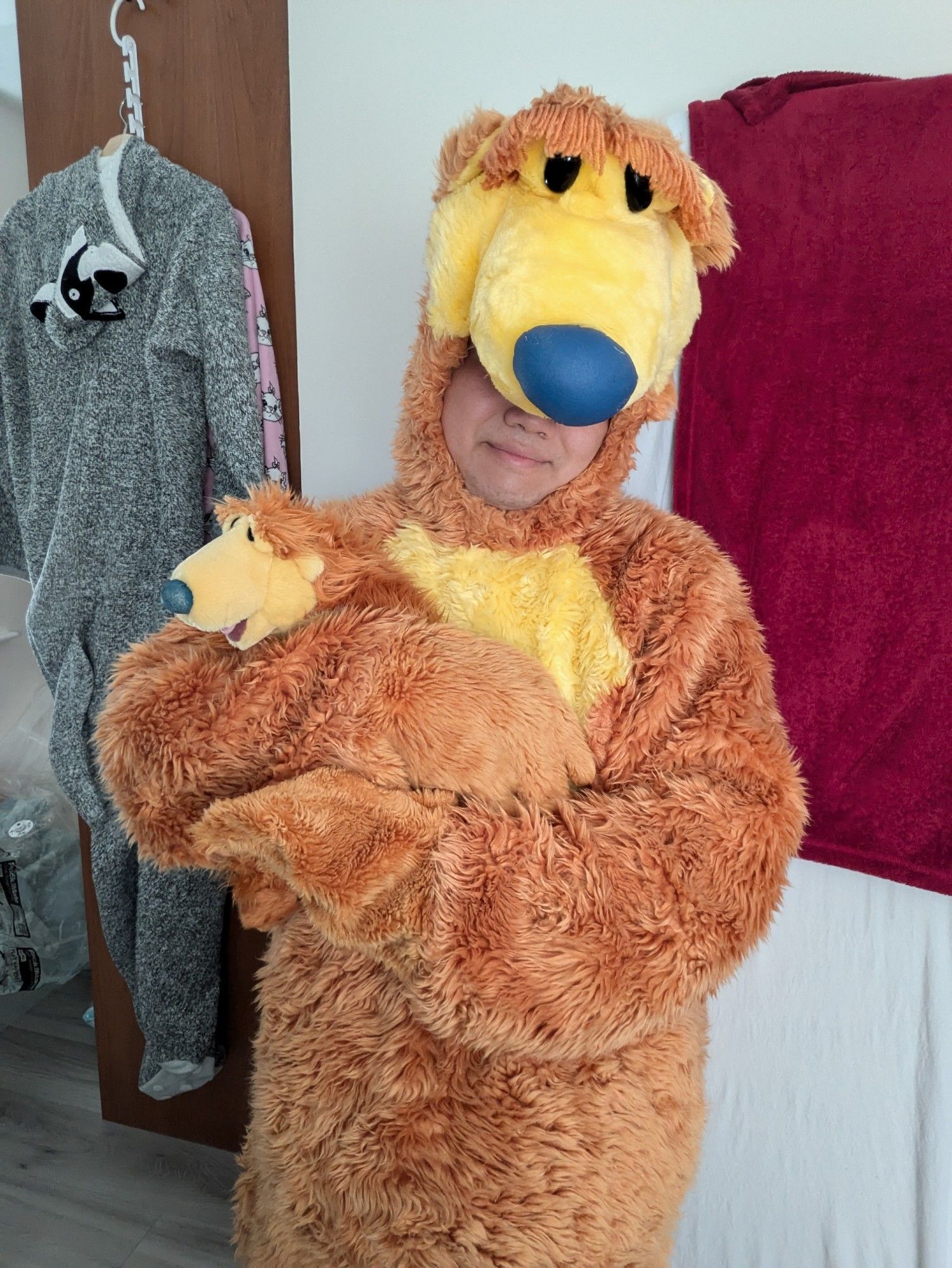 A person wearing a Bear in the Big Blue House costume is standing in a room while hugging a small Bear in the Big Blue House plush toy.