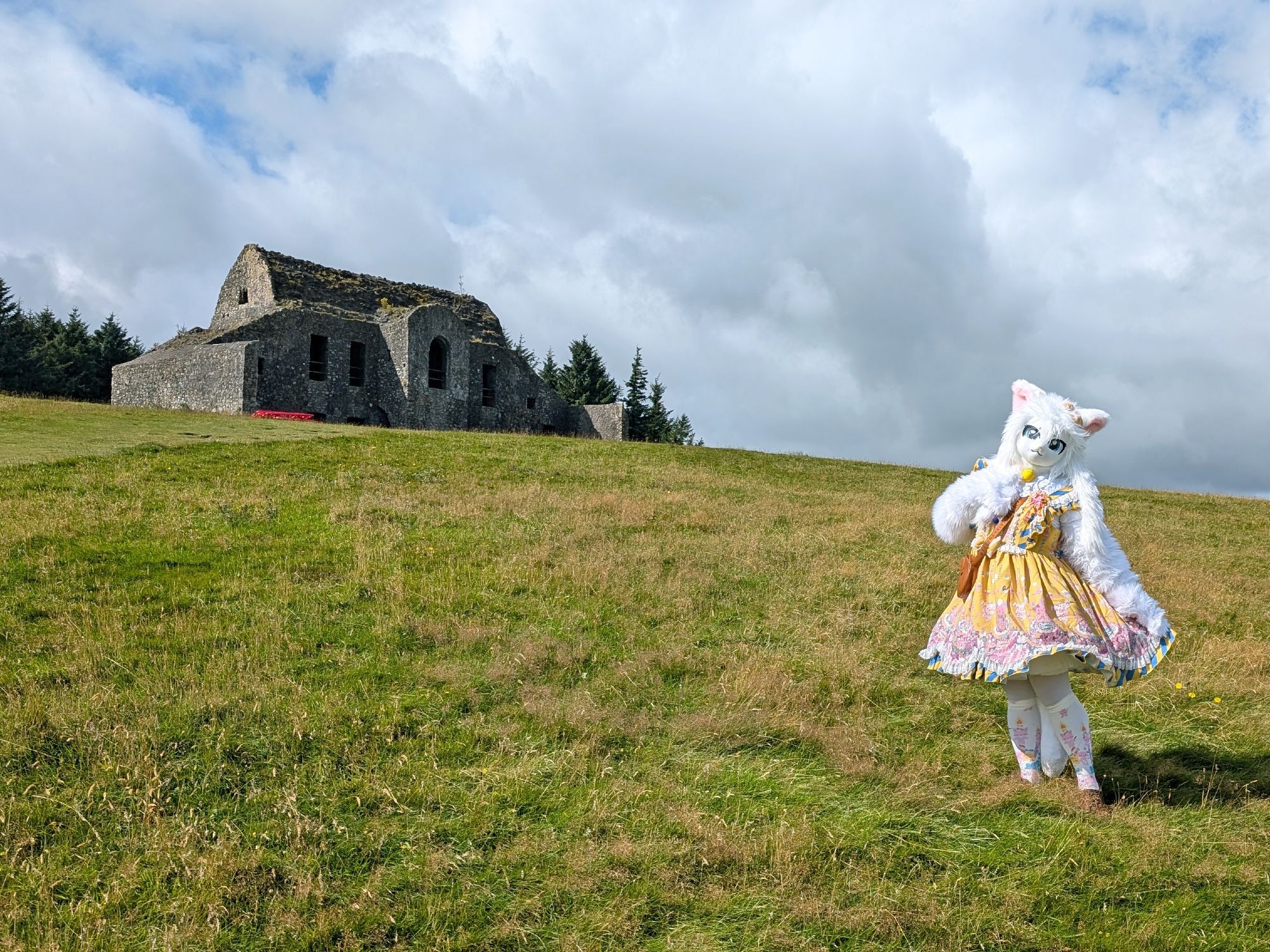 A white cat fursuiter with a yellow dress is standing is a grass field with a pose. Behind them in a distance is an old stone building.