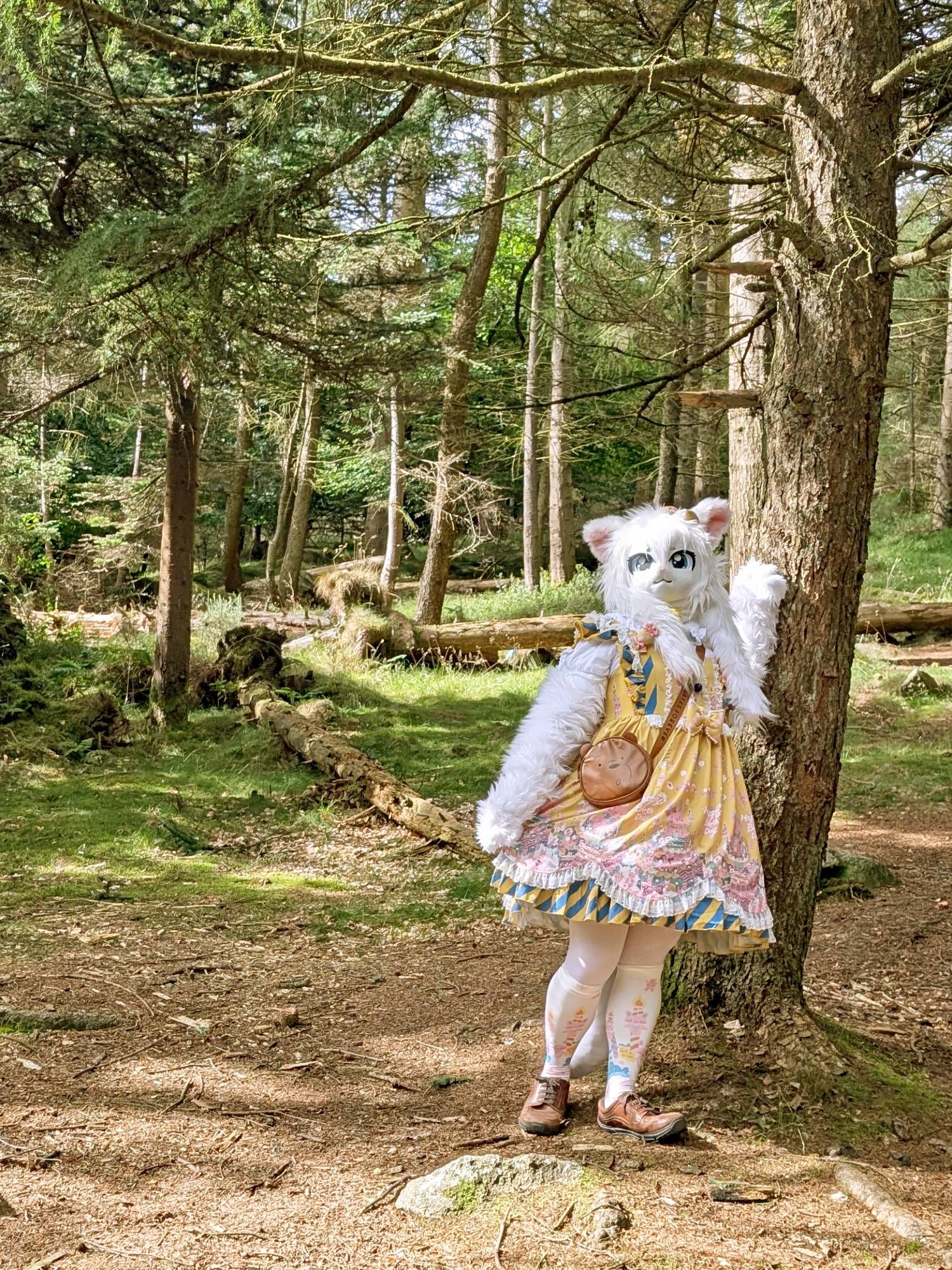 A white cat fursuiter with a yellow dress is standing next to a tree with their left hand on the tree trunk and they're looking up to their right.