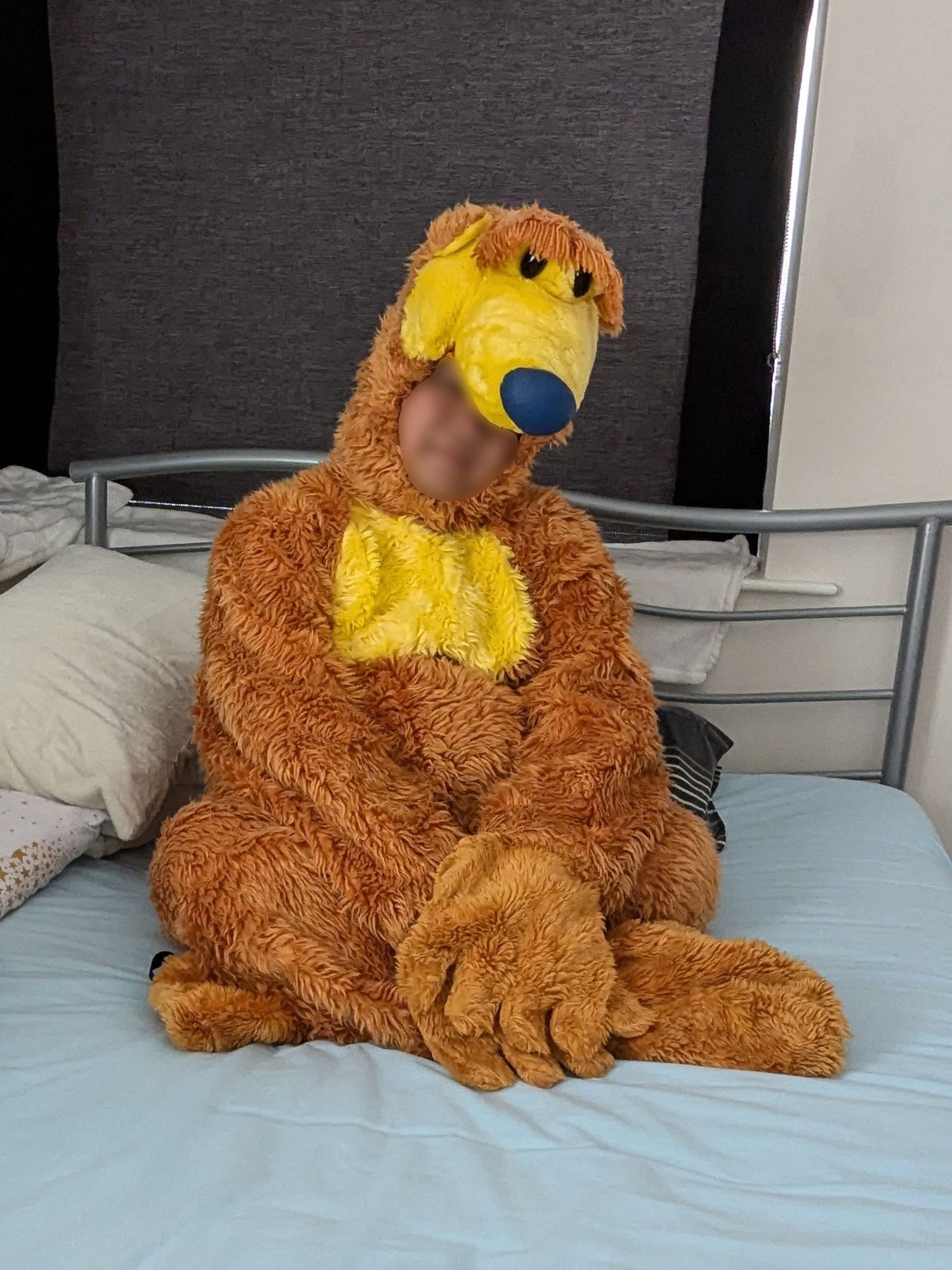 A person with blurred face is sitting on a blue bed wearing a Baer in the Big Blue House costume.