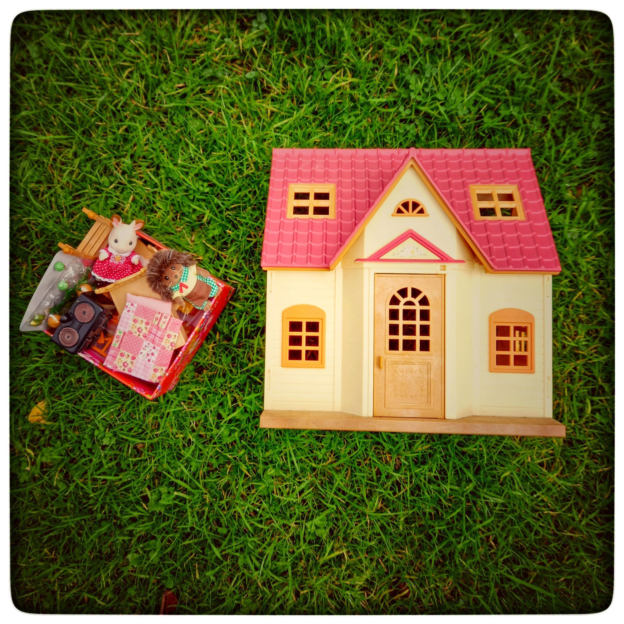 A small plastic dollhouse & a box of jumbled furniture & sylvanian creatures, all on green grass.