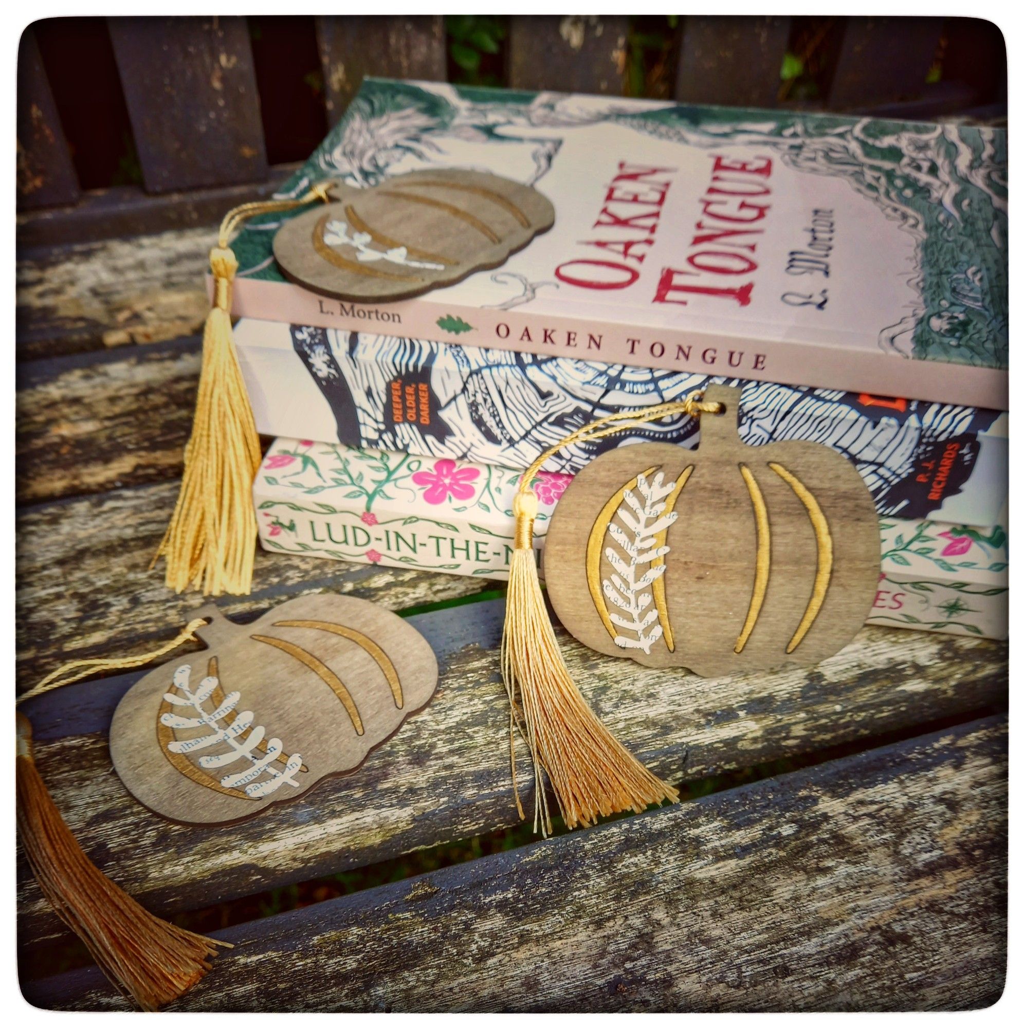 On an aged garden bench with greenery behind is a stack of books - Oaken Tongue by L. Morton, Deeper Older Darker by P.J Richards, & Lud in the Mist by Hope Mirrlees - with wooden pumpkin bookmarks scattered about with fancy gold details & book page botanicals & gold tassels.