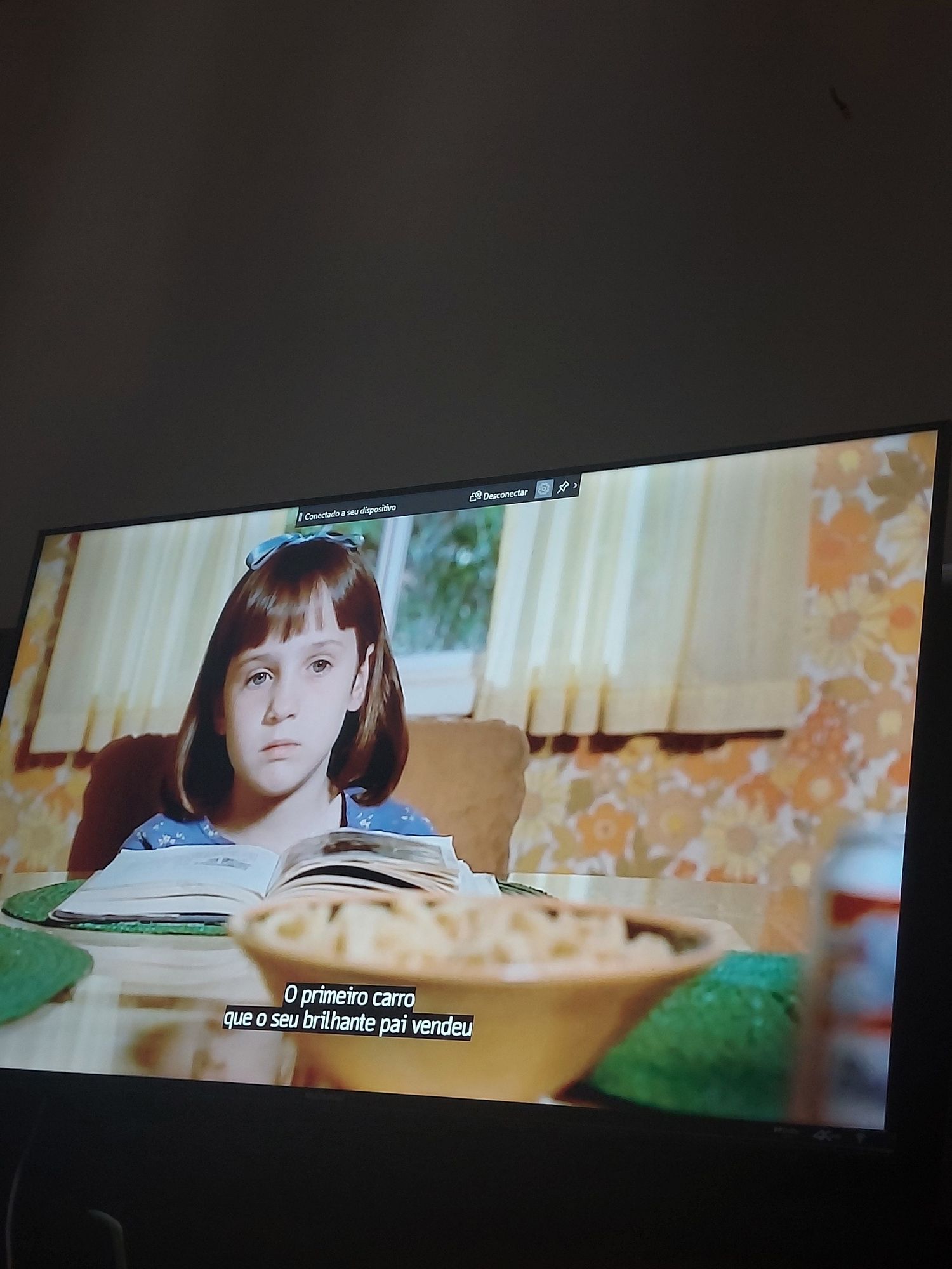 Foto de uma TV exibindo o filme Matilda, na tela está uma menina branca de cabelos lisos curtos e franja, ela usa um laço azul no cabelo e está com o rosto sério com um livro em mãos. Na legenda está escrito 'o primeiro carro que o seu brilhante pai vendeu'