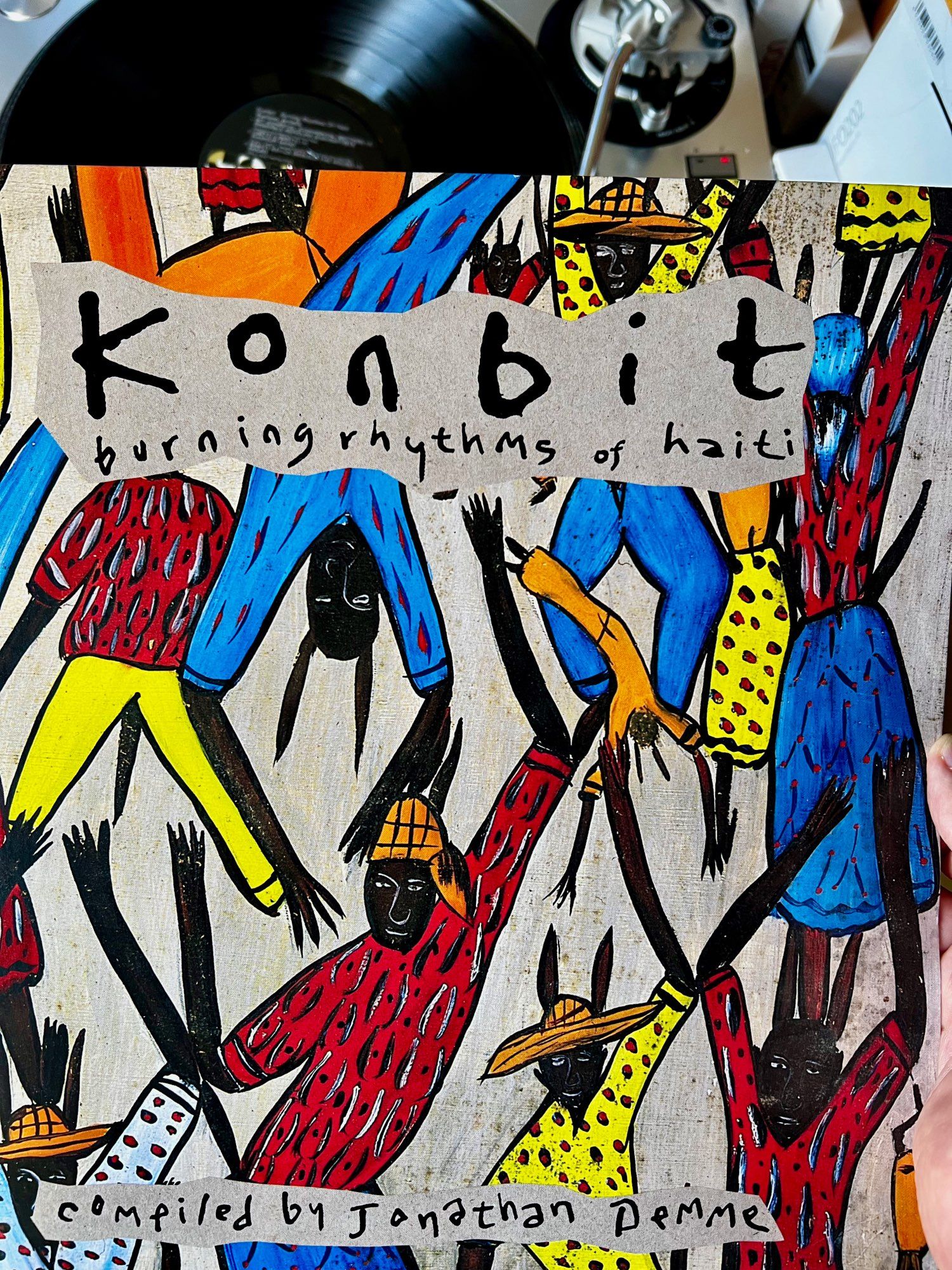 An album cover titled "Konbit: Burning Rhythms of Haiti," compiled by Jonathan Demme. The artwork features colorful, abstract figures dancing and celebrating, with vibrant clothing and expressive poses. A vinyl record player is partially visible in the background.