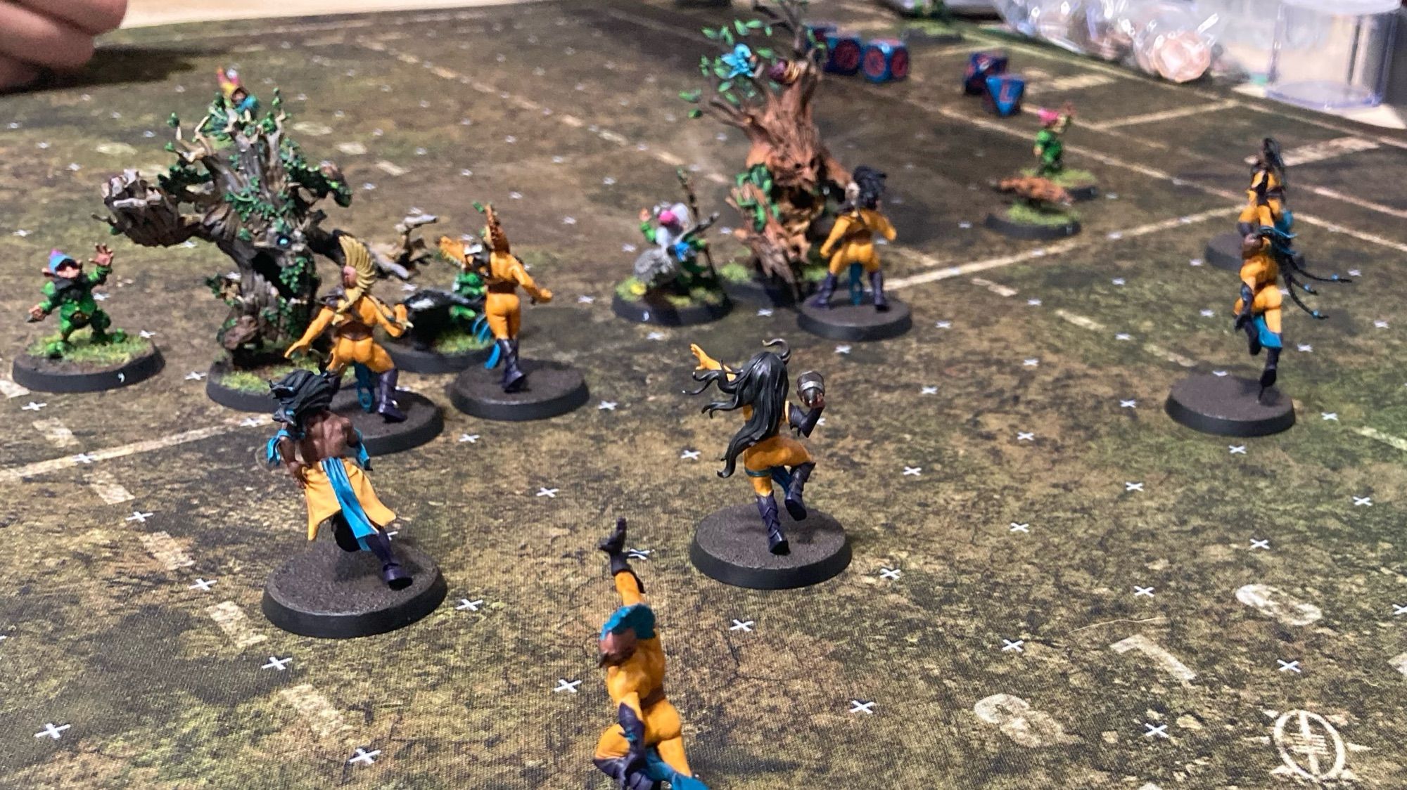 A Blood Bowl pitch depicting a grassy field with squares demarcated on it. Two teams are present: Colorful gnomes in blue green and red with two treemen, and wood elves in yellow and blue