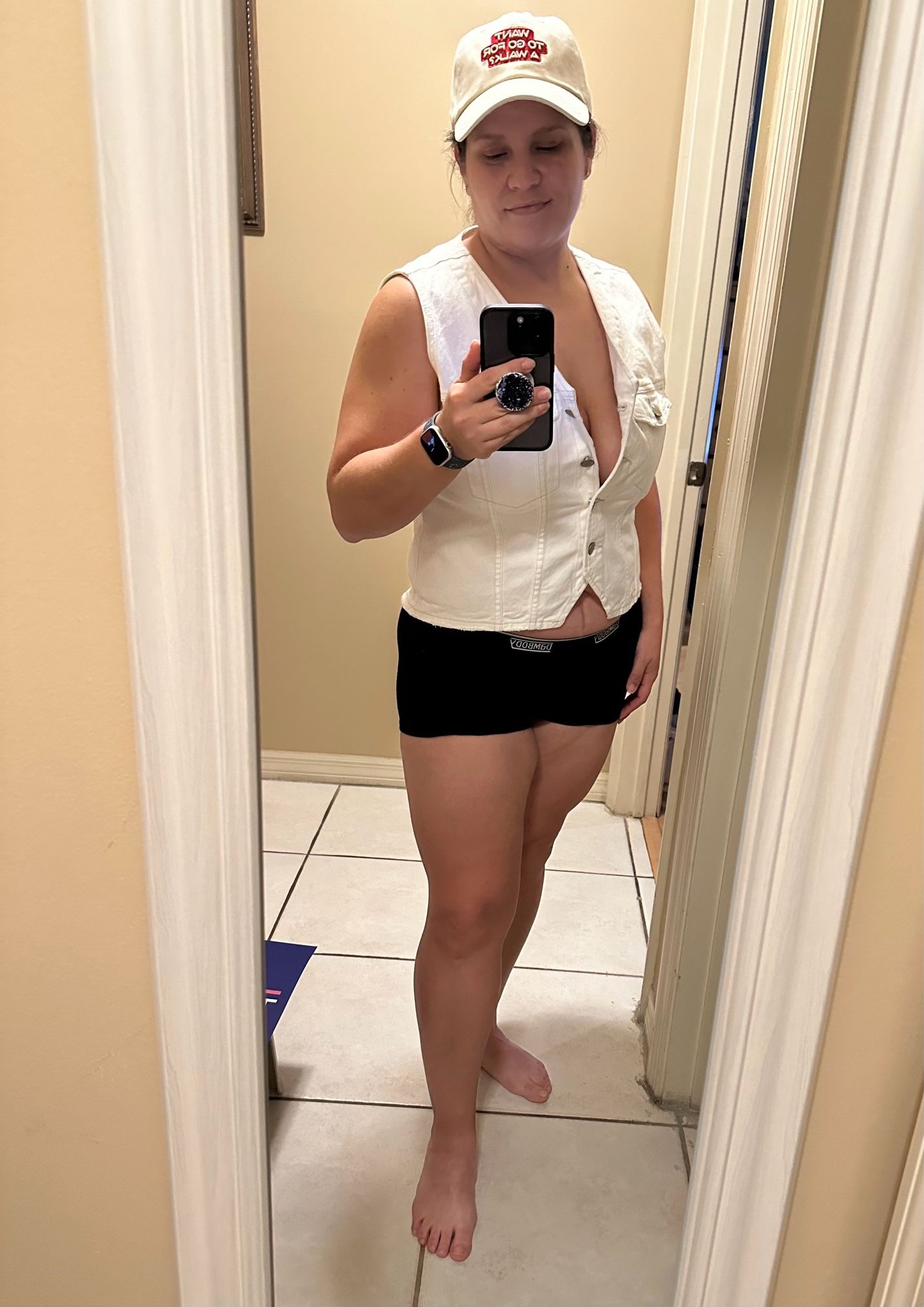 Posing for a selfie in front of a mirror in short black shorts and a white denim vest opened slightly. My white hat says “want to go for a walk?” which is now my favorite thing