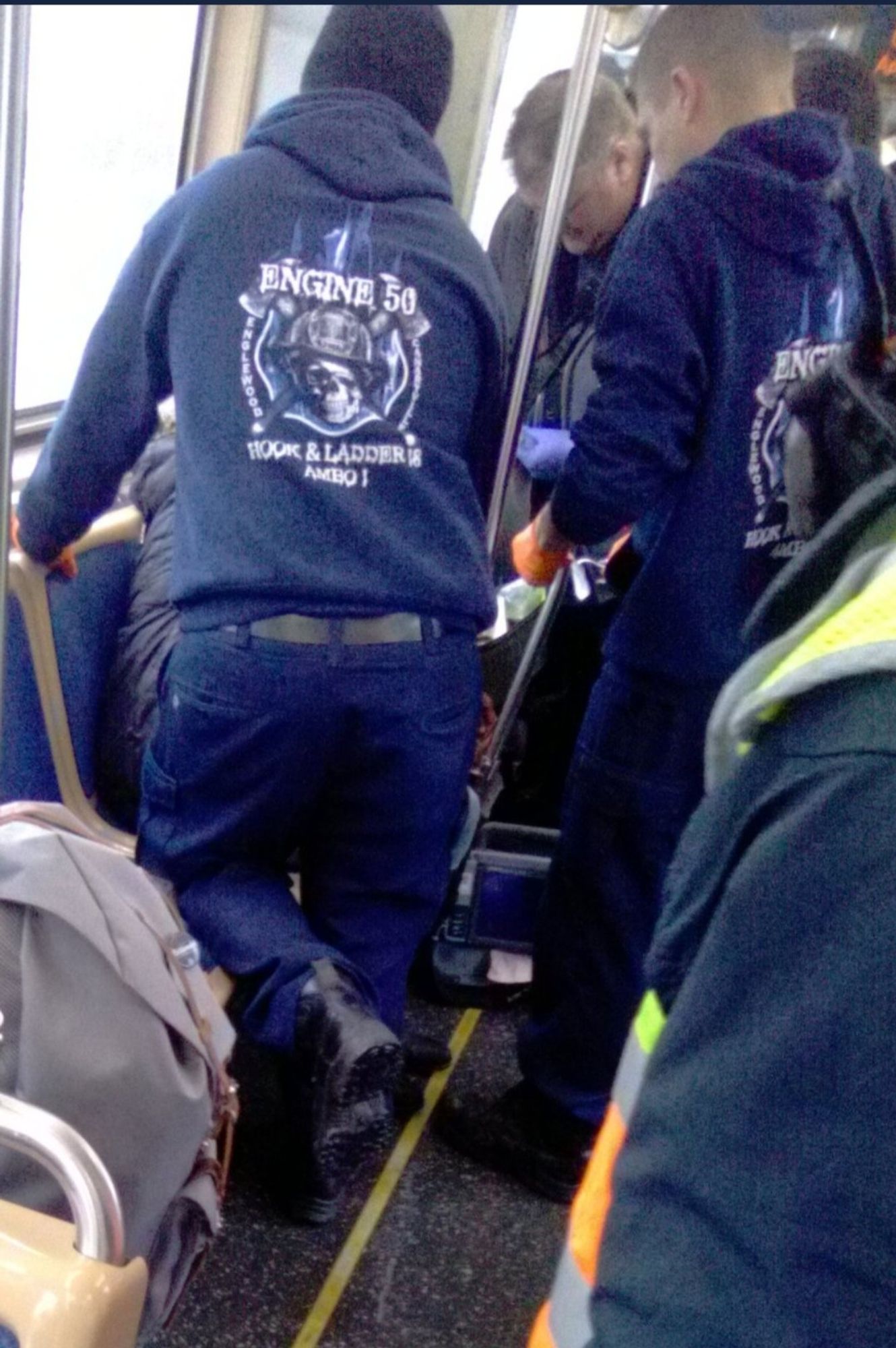 Three paramedics in matching navy hoodies respond to a medical emergency on a CTA bus. The hoodies say "engine 50 hook & ladder Ambo 1 Englewood Canaryville" and depict a skull and crossbones type design, but the crossbones are axes and the skull is wearing a firefighter's hat