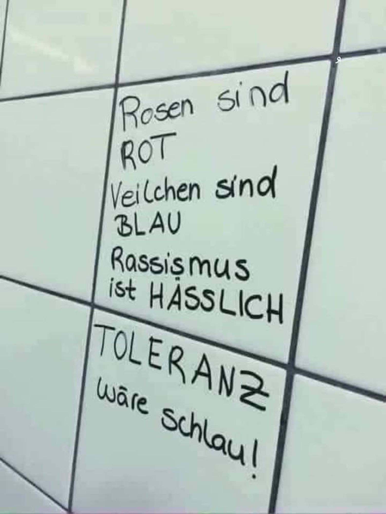 Weiße Fliesen mit dunklen Fugen, in schwarzer Schrift steht auf den Fliesen: Rosen sind ROT, Veilchen sind BLAU, Rassismus ist HÄSSLICH, TOLERANZ wäre schlau!