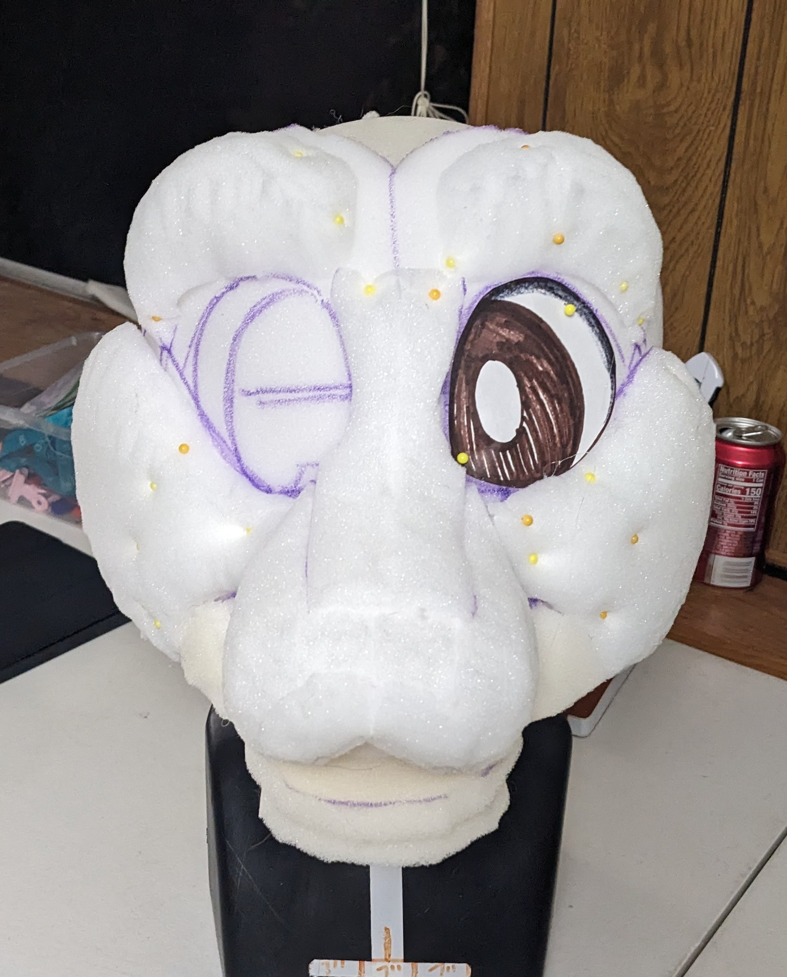 Front view of a white upholstery foam fursuit head base with one brown paper eye and lots of yellow pins holding the foam in place