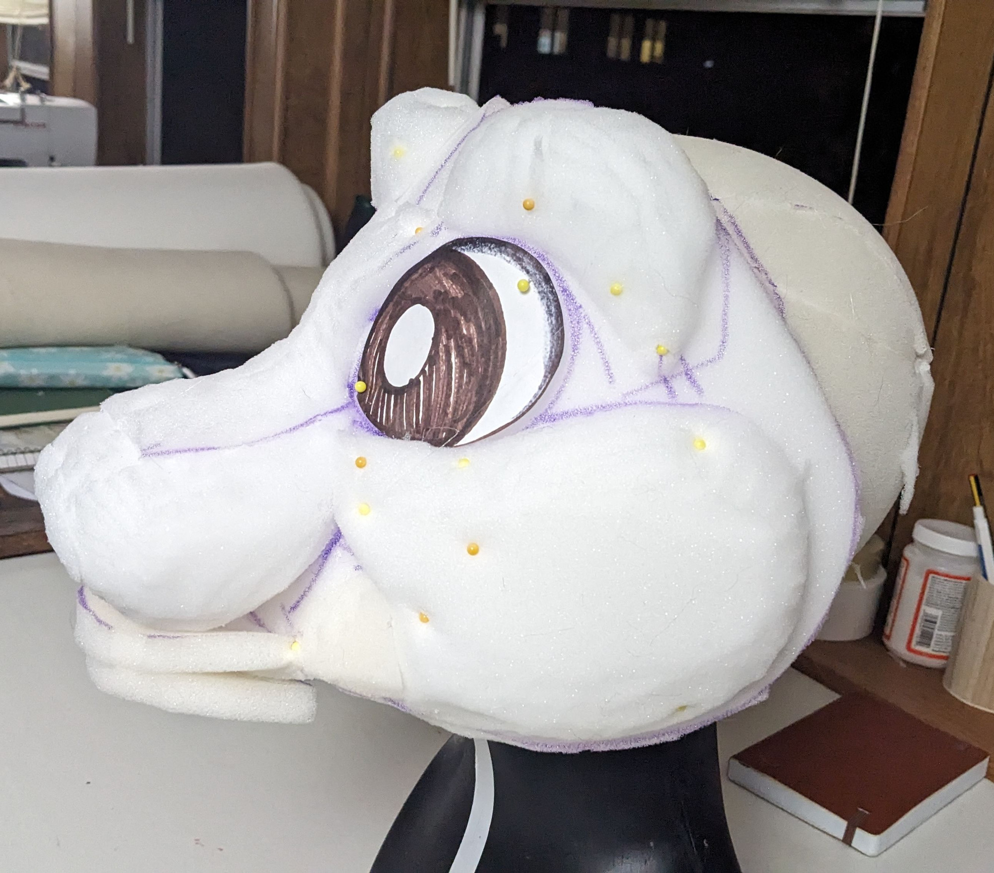 Side view of a white upholstery foam fursuit head base with one brown paper eye and lots of yellow pins holding the foam in place