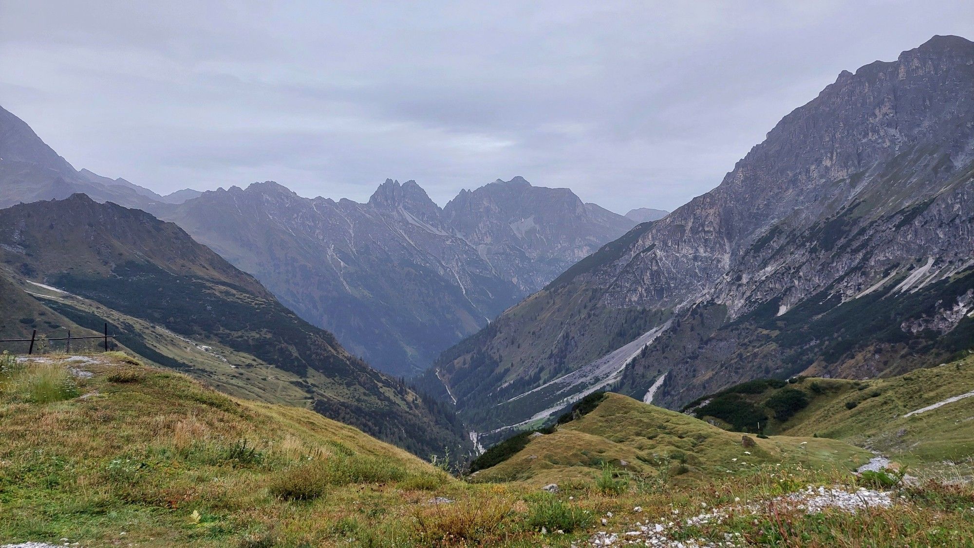 Blick von einer Bergwiese auf hohe Felsberge. Komplett bewölkt, regnerisch.