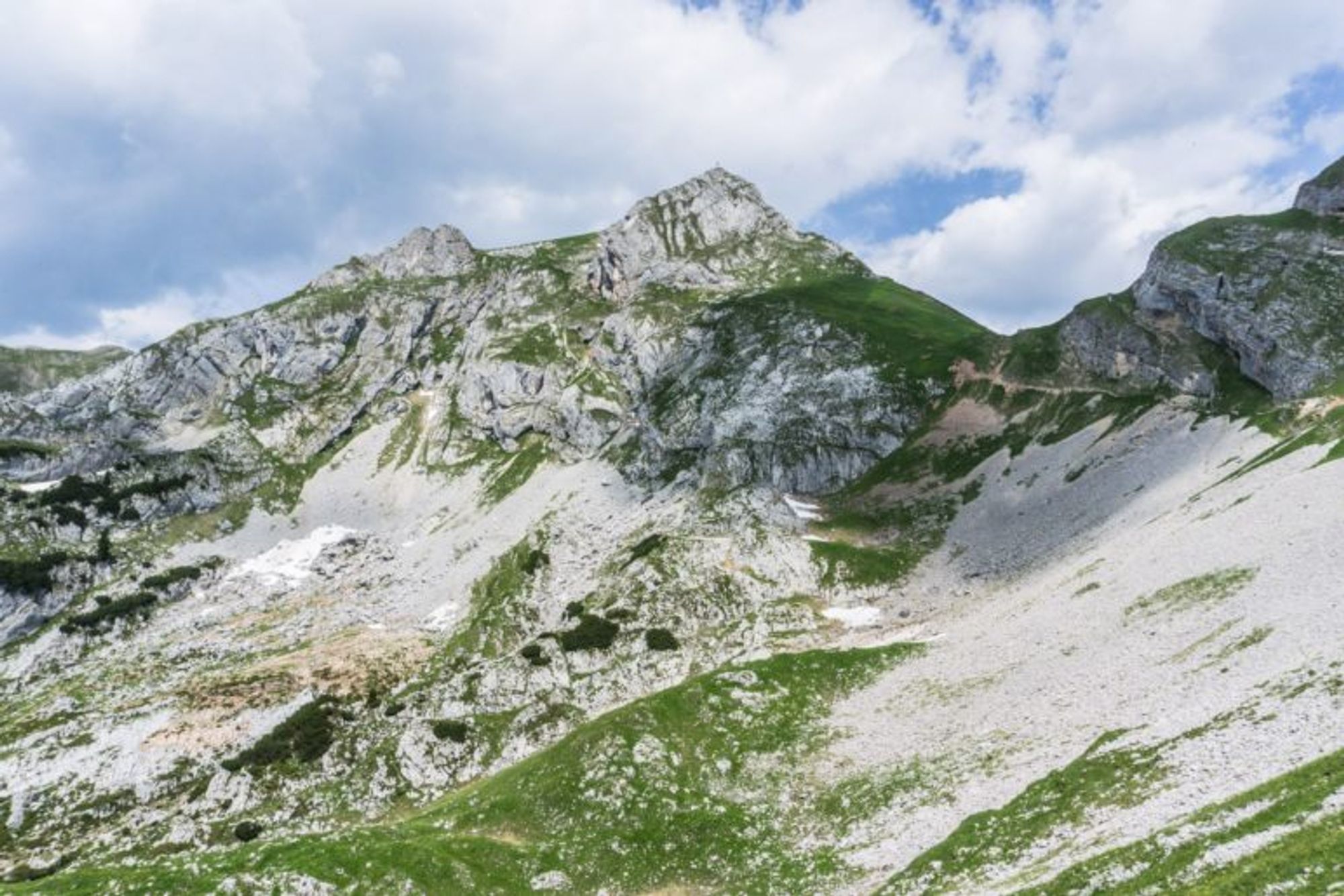 Blick auf die Hochiss, ein Berg mit pyramidenförmiger Felsspitze. Unterhalb ist das Gelände gemischt felsig und mit Wiesen durchsetzt. Ein sonniger, leicht bewölkter Tag.