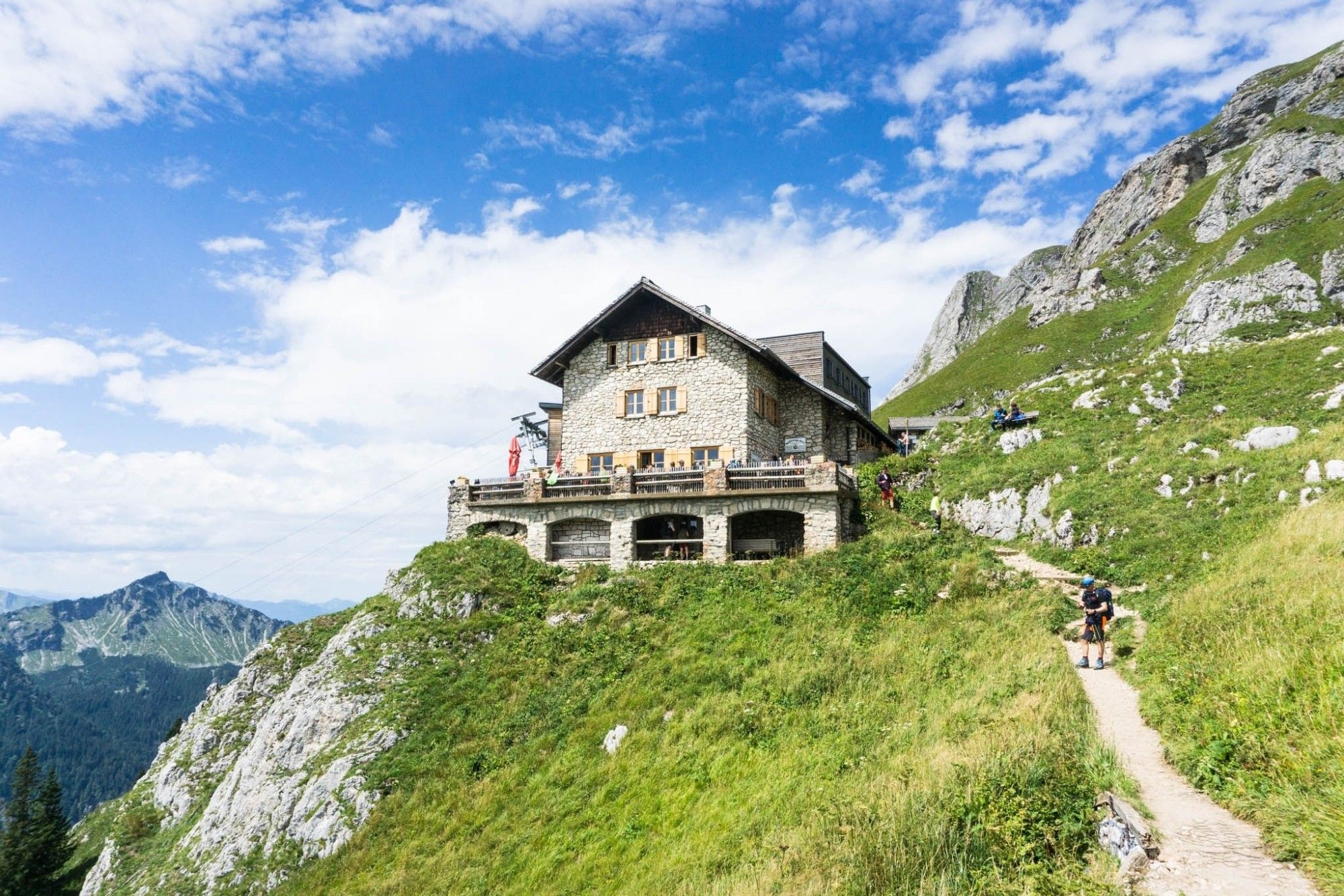 Eine massive Berghütte aus grauem Stein. Vor der Hütte eine Terrasse. Die Hütte steht auf einem Vorsprung auf einer Wiese. Im Hintergrund grüne Bergflanken.