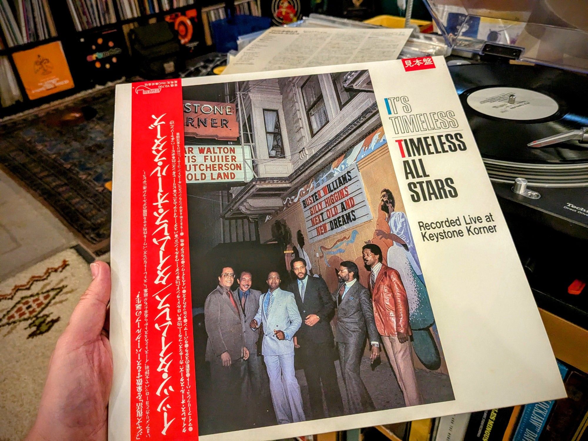 front cover of "It's Timeless: Timeless All Stars Recorded Live at Keystone Korner" lp with a red obi-strip, held in hand in front of the whitelabel promo playing. cover shows the six members standing in front of the club under the sign announcing them. - vinyl record wall in the background.