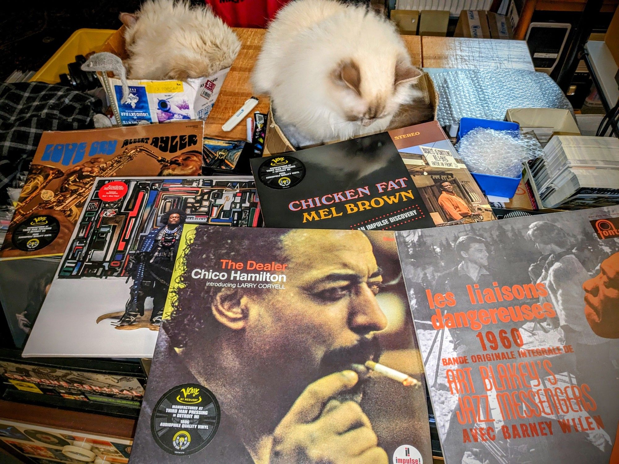 five still sealed jazz vinyl records laid out over the Technics, most prominent Chico Hamilton "The Dealer" smoking. On the packing table behind them are cats Emil sleeping and Milo inspecting the record hanging over his cardboard box.