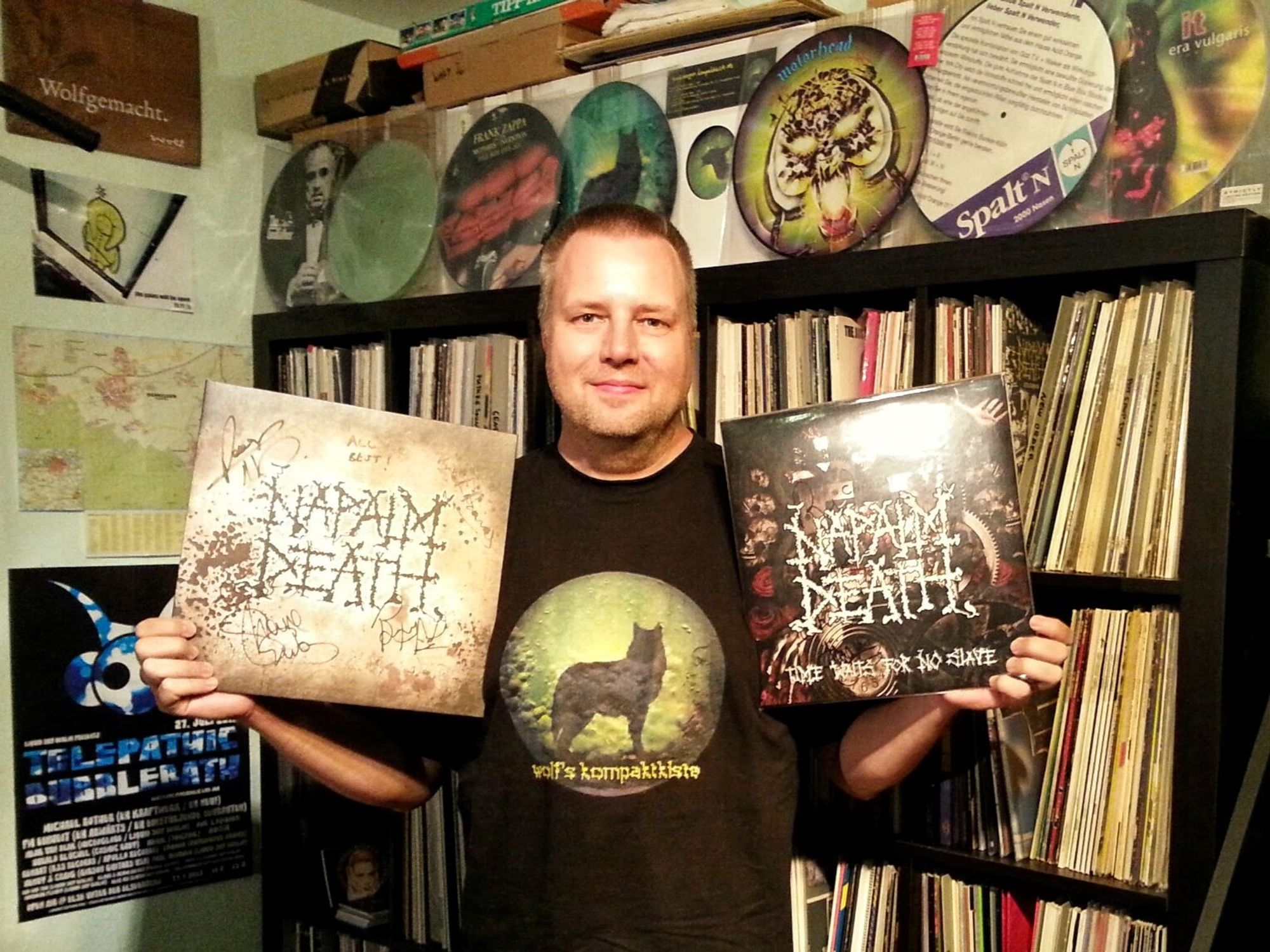 wolf @ kompaktkiste holding up two signed versions of the Napalm Death LP "Time waits for no slave" in front of a vinyl record collection wall with some picture discs on top