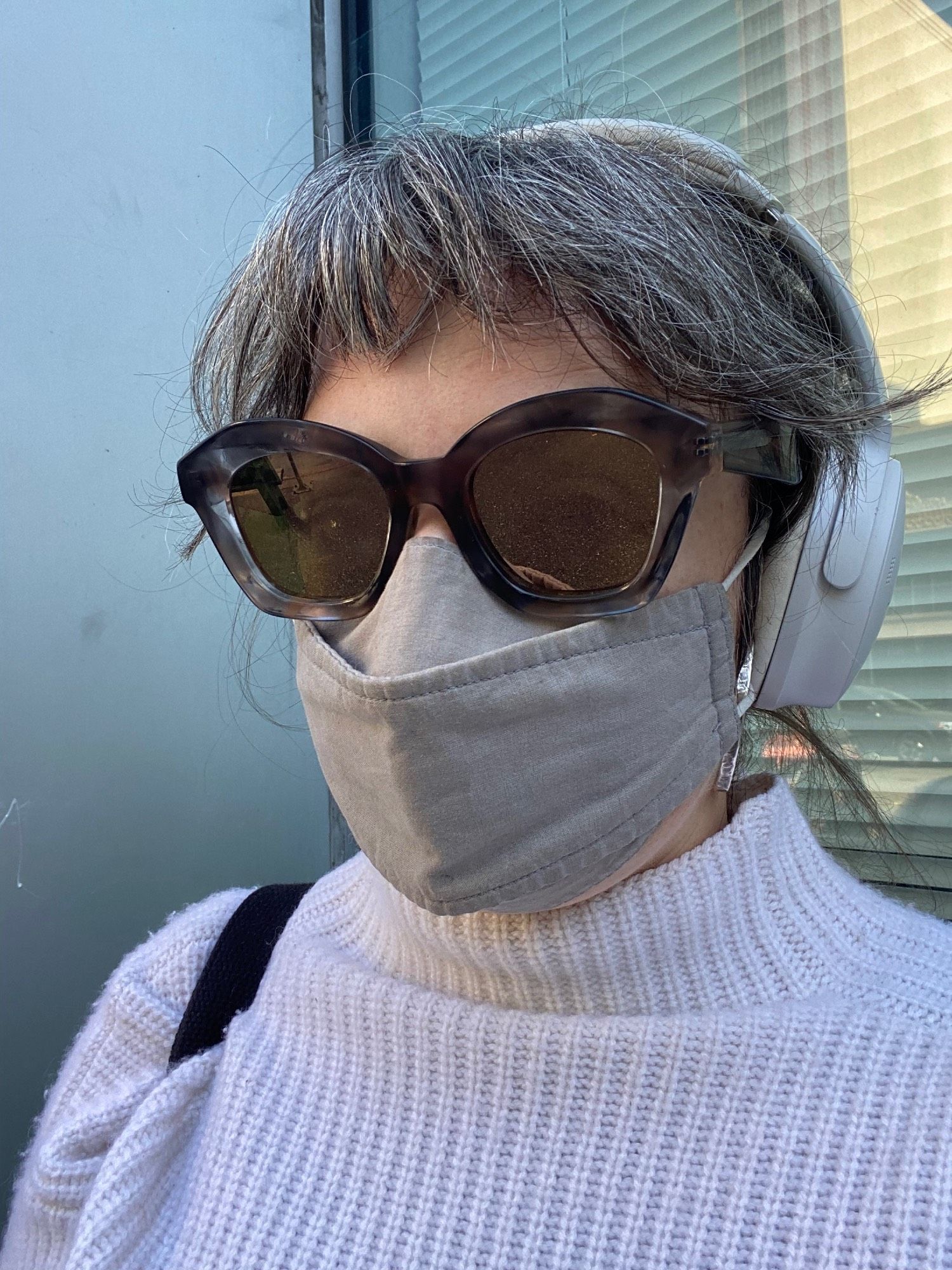 a person wearing a white sweater, stiff gray opaque face mask covering their entire chin, mouth, and nose coming up above the bridge, large sunglasses with mirrored lenses covering the entire eye area including eyebrows and overlapping the face mask, and bangs covering the forehead, plus large over-ear headphones, leaving the viewer to wonder how any kind of “look” might be conveyed through these myriad physical barriers much less a “dirty” one