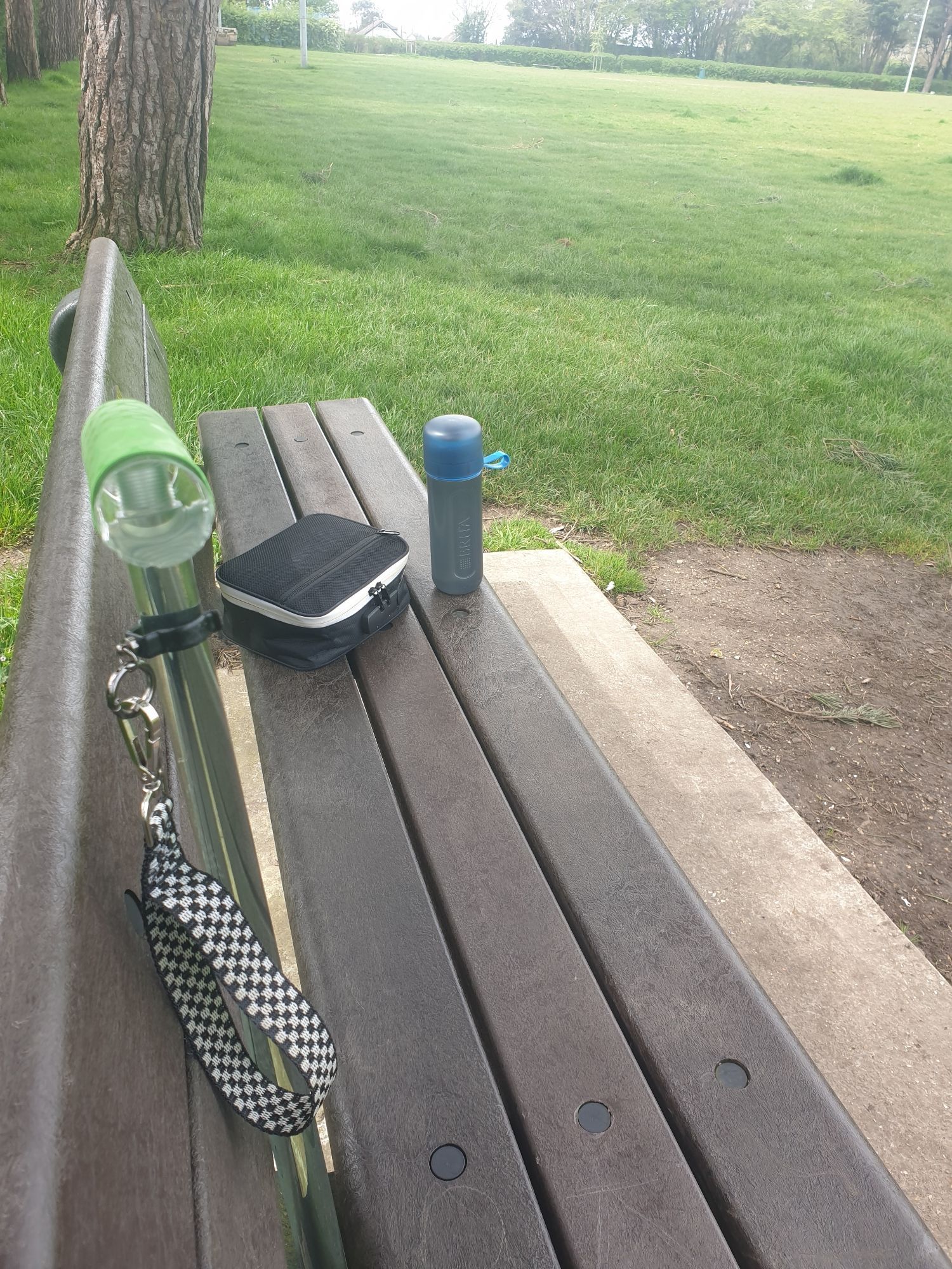 A park bench with a walking stick leant against it , a water bottle and a smellproof locked bag