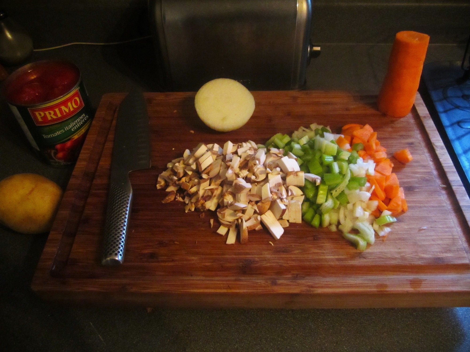 A photo of diced carrots, onion, celery & mushrooms on a cutting board ready to become soup stock. An open can of Roma tomatoes & a potato are nearby.