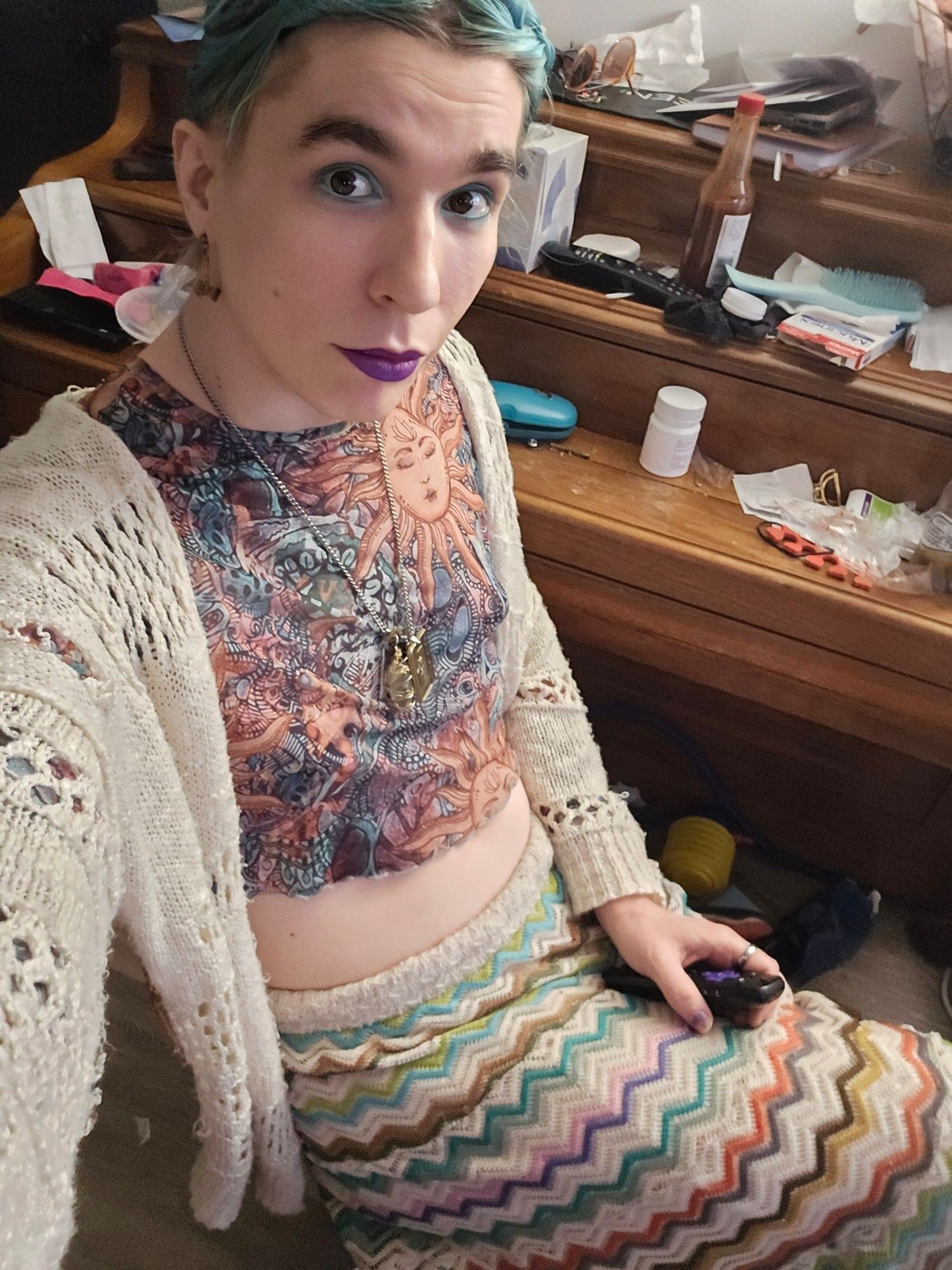 The image depicts a person sitting in what appears to be a cozy, slightly cluttered room. The individual is dressed in a colorful, patterned outfit consisting of a tight-fitting crop top with artistic designs incorporating a sun motif, and a long skirt with a zigzag pattern in various bright colors. They are wearing a crocheted, cream-colored cardigan over the top. They have blue-green hair styled in a braid and wear bold makeup, including purple lipstick and blue eyeshadow. The person is holding a black video game controller in one hand.

In the background, there is a wooden piano or desk covered with various items including a hot sauce bottle, sunglasses, a hairbrush, a container of pills, and some papers, giving an impression of a lived-in space.