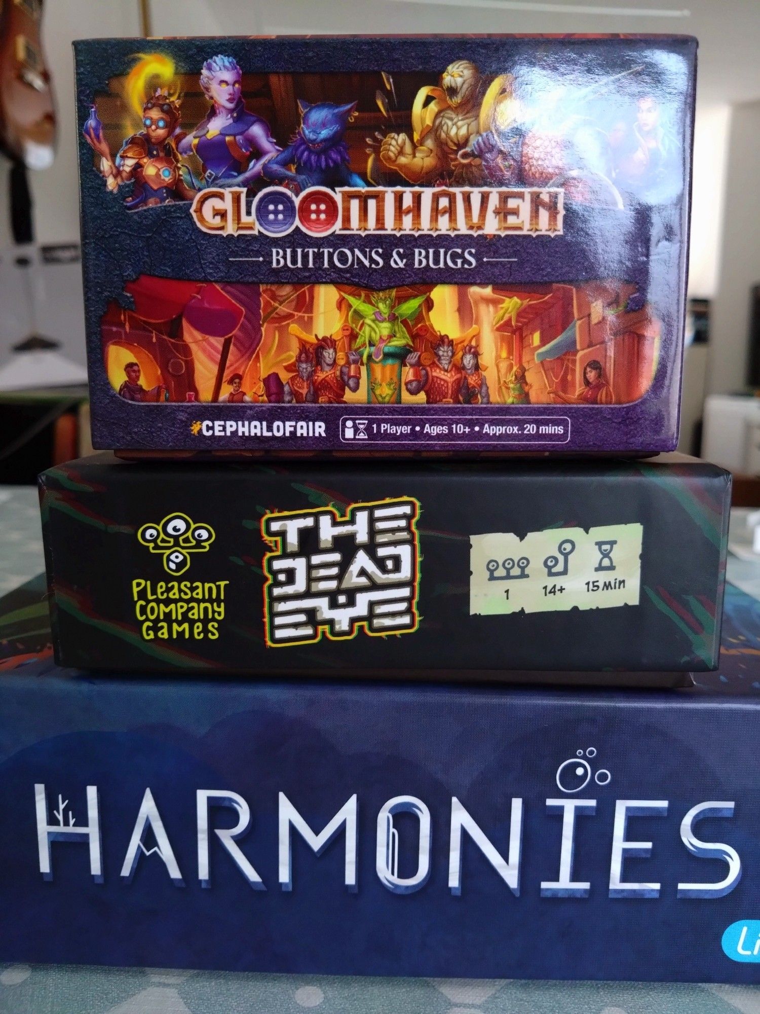 A pile of boardgames, from top to bottom: Gloomhaven Buttons&Bugs, The Dead Eye and Harmonies