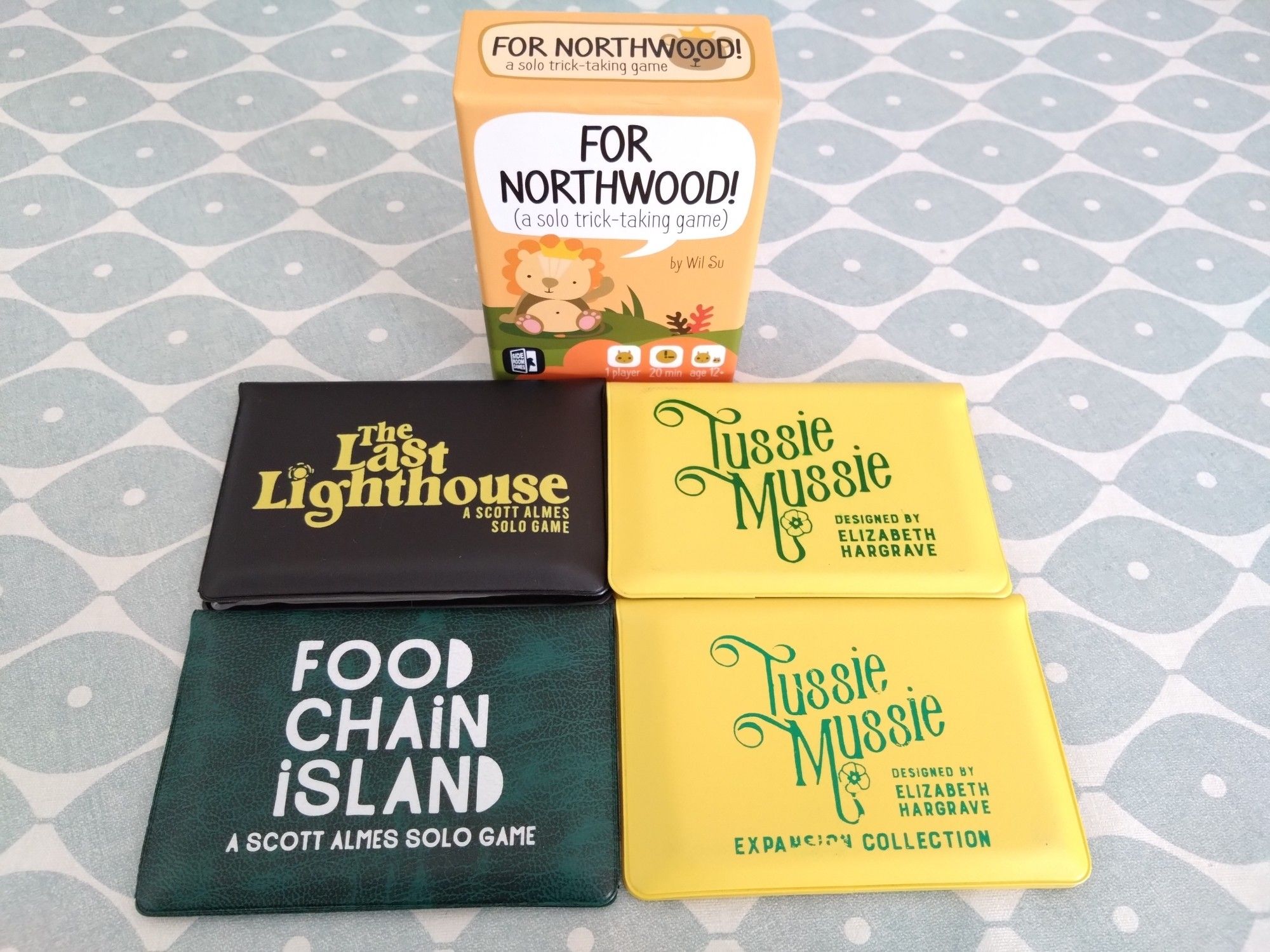 Four boardgames are pictured: For Northwood! in a small box, and the three games in wallets are The Last Lighthouse, Food Chain Island and Tussie Mussie with the expansion collection.