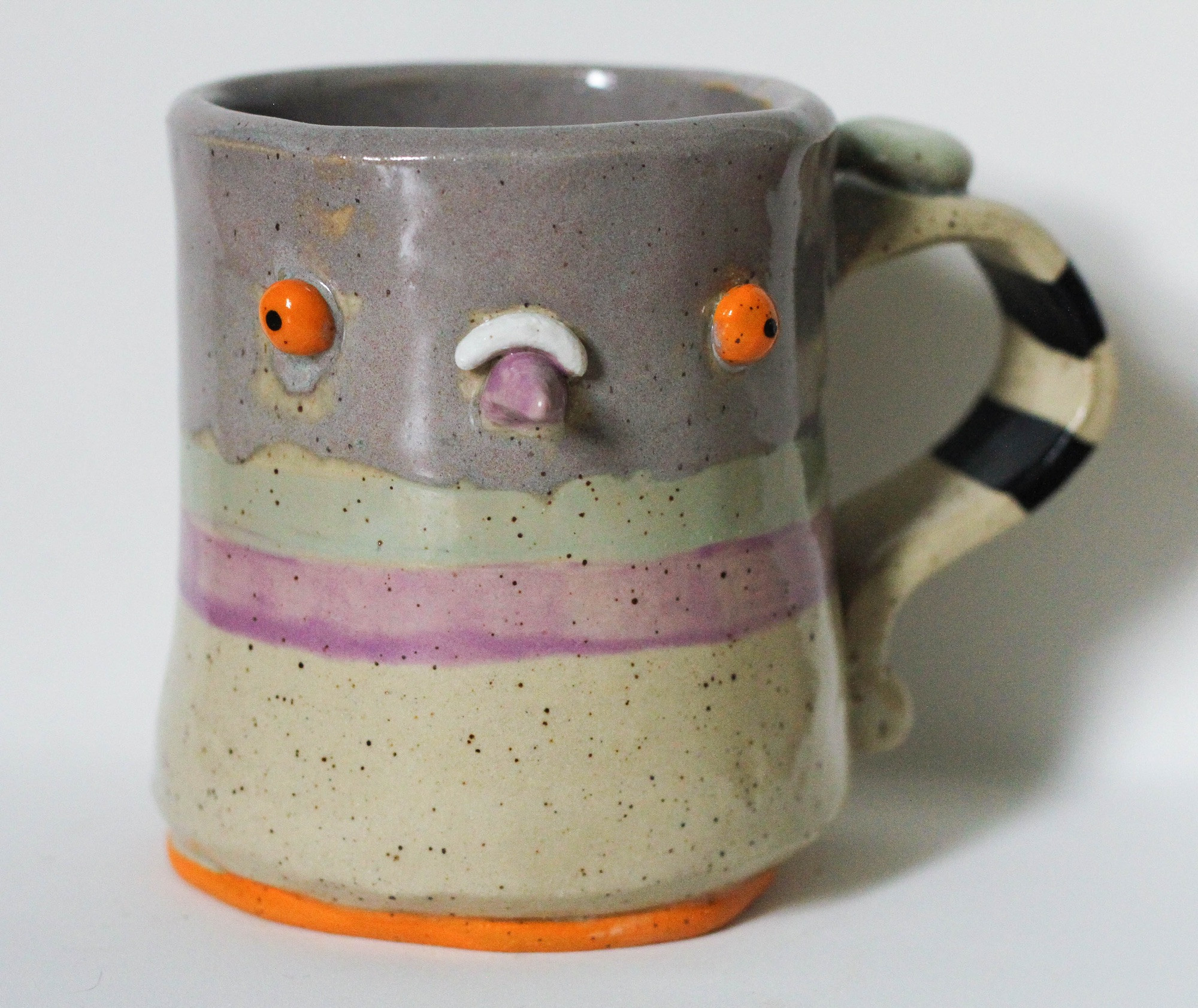 a light speckled mug with the top half glazed grey, and two stripes directly below (mint and lilac). the handle has two black stripes around it. the foot is painted bright orange and there are two bright orange eyes as well as a little purple beak. PIGEON MUG BABY