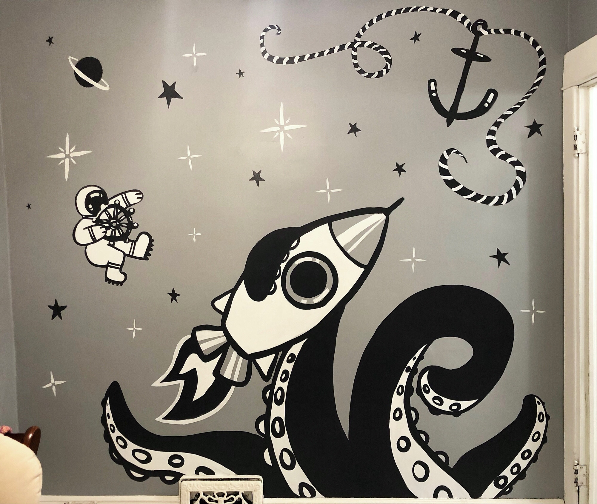 a grey wall with a black and white mural of big cartoony sea monster tentacles grasping a cartoon spaceship. an astronaut holds an old ship wheel and an anchor with a rope float among the stars and planets