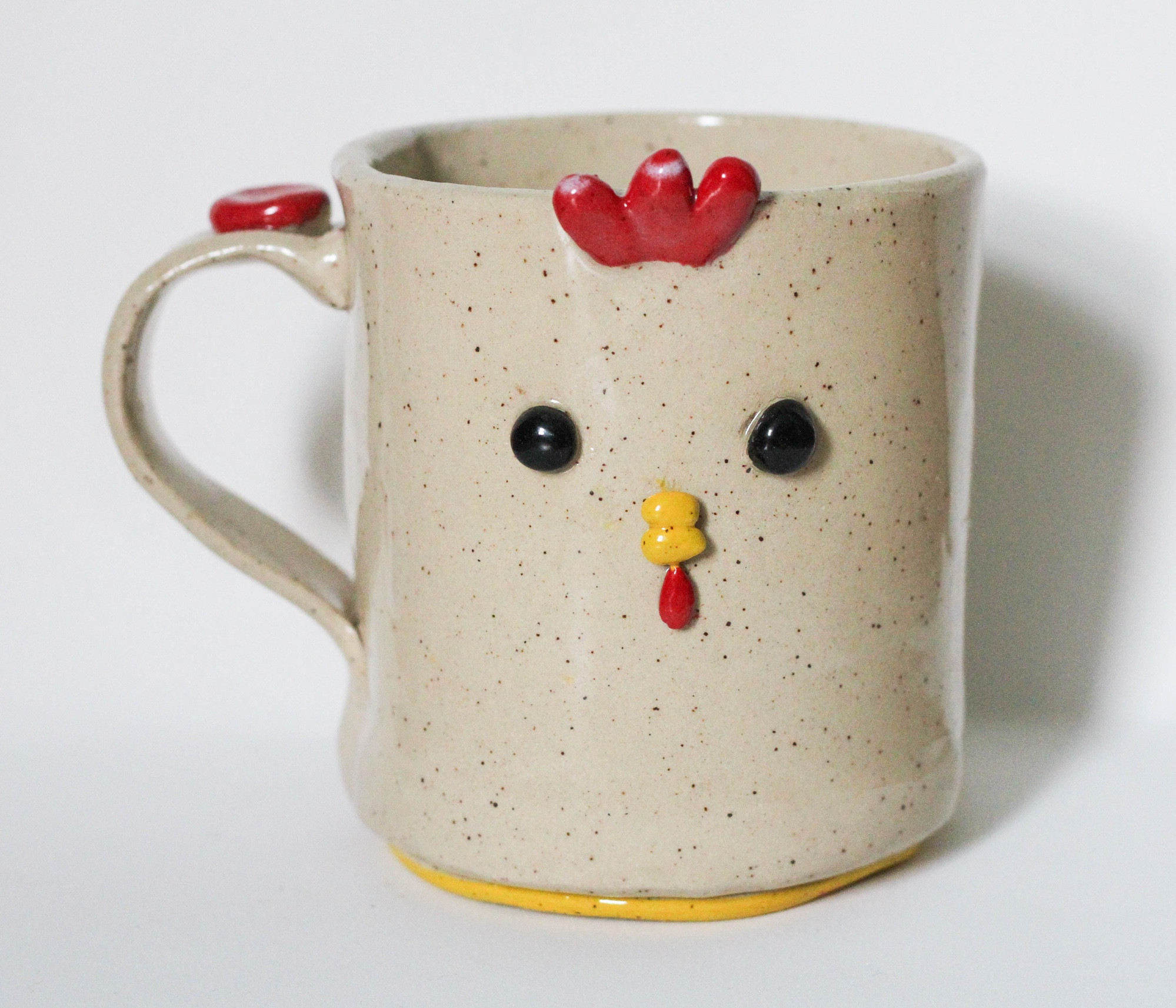 a big mug with light speckled clay. the foot is painted yellow. there is a little yellow beak, a couple beady black eyes, and a red… wattle? i don’t remember what all that’s called but it’s a CHICKEN a little chicky mug i loved her but she sold so fast