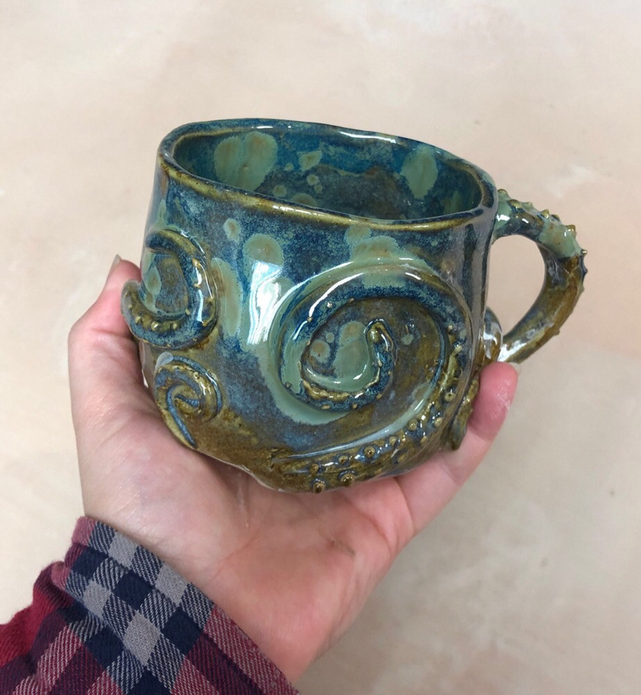 a mug with blue and green splotchy shipwreck colours. tentacles wrap around it and the handle is also a tentacle 