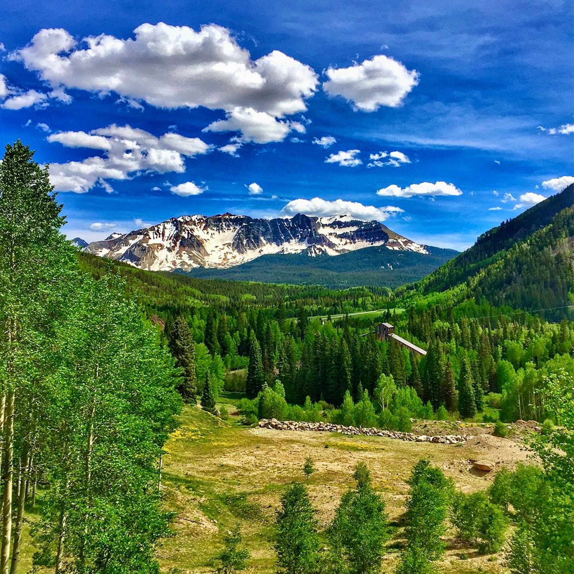 Color photo of a wide meadow with a saw mill at the back and tall snow capped mountains in the distance.